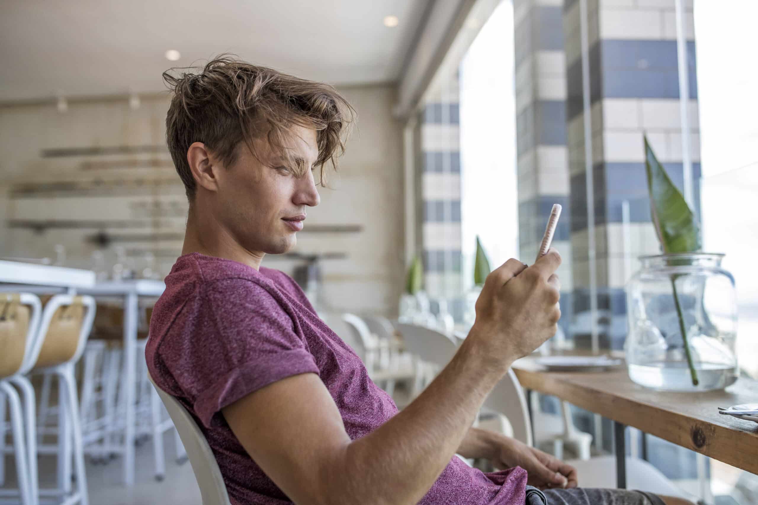 Ein junger Mann in einem lässigen T-Shirt sitzt an einem Cafétisch und konzentriert sich auf sein Smartphone. Das moderne Interieur und die großen Fenster suggerieren eine helle, gesellige Atmosphäre. © Fotografie Tomas Rodriguez