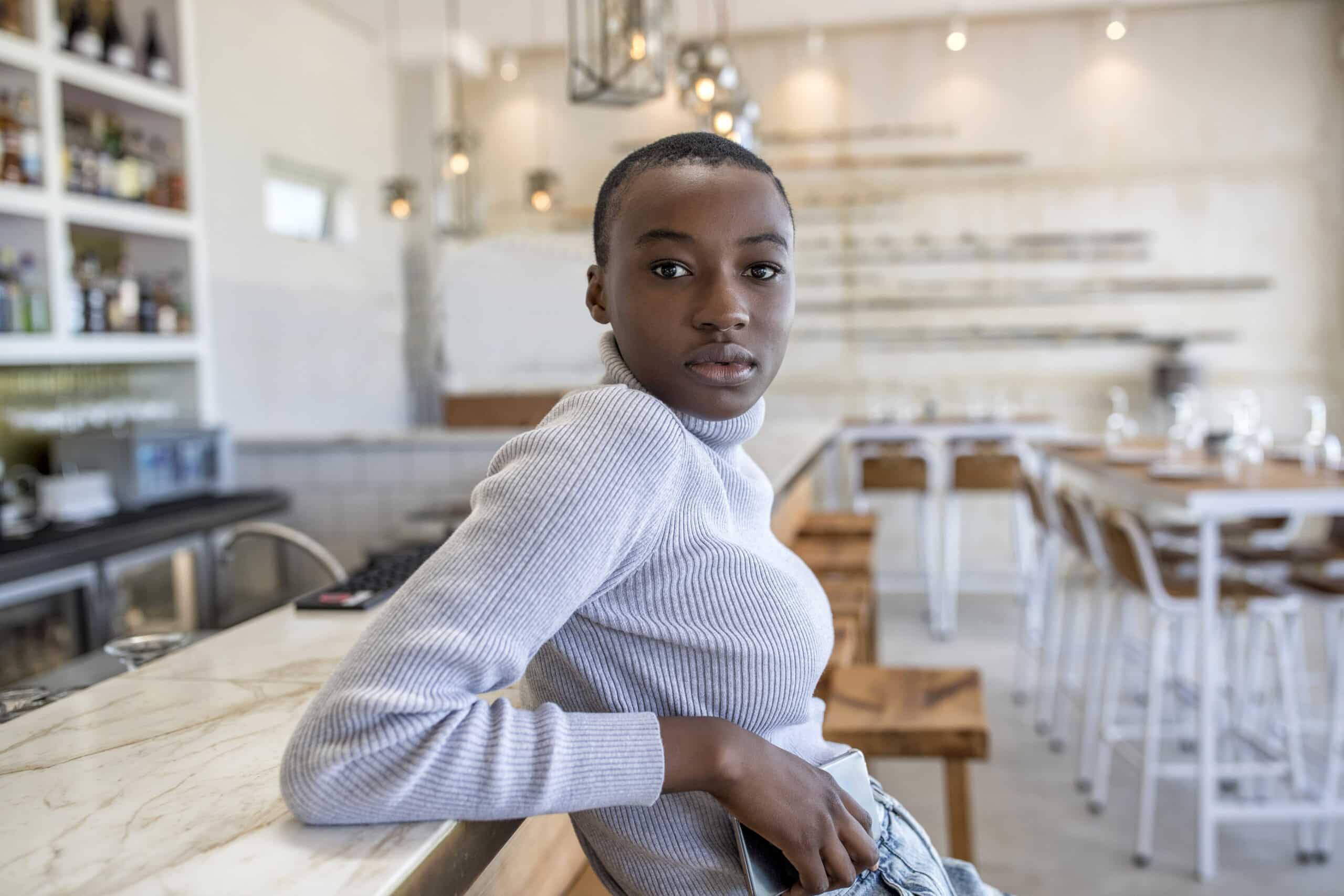 Eine junge schwarze Frau in einem weißen Rollkragenpullover sitzt an einer Marmortheke in einem hellen, modernen Café und blickt mit ruhigem Gesichtsausdruck direkt in die Kamera. © Fotografie Tomas Rodriguez