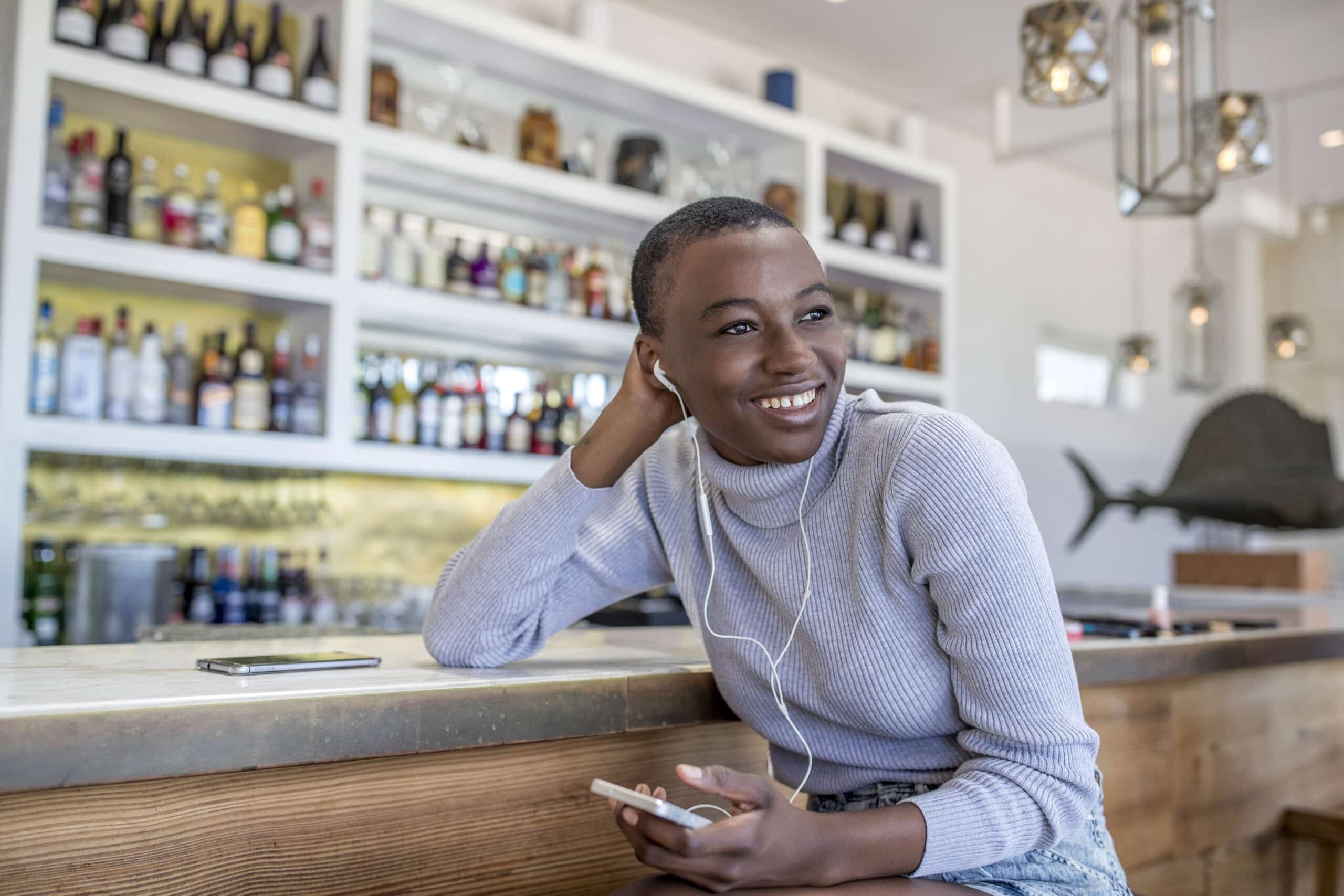 Eine fröhliche junge Frau mit Kurzhaarschnitt und hellgrauem Pullover sitzt an einer mit verschiedenen Weinflaschen aufgereihten Bar, benutzt ihr Smartphone und trägt Kopfhörer. © Fotografie Tomas Rodriguez