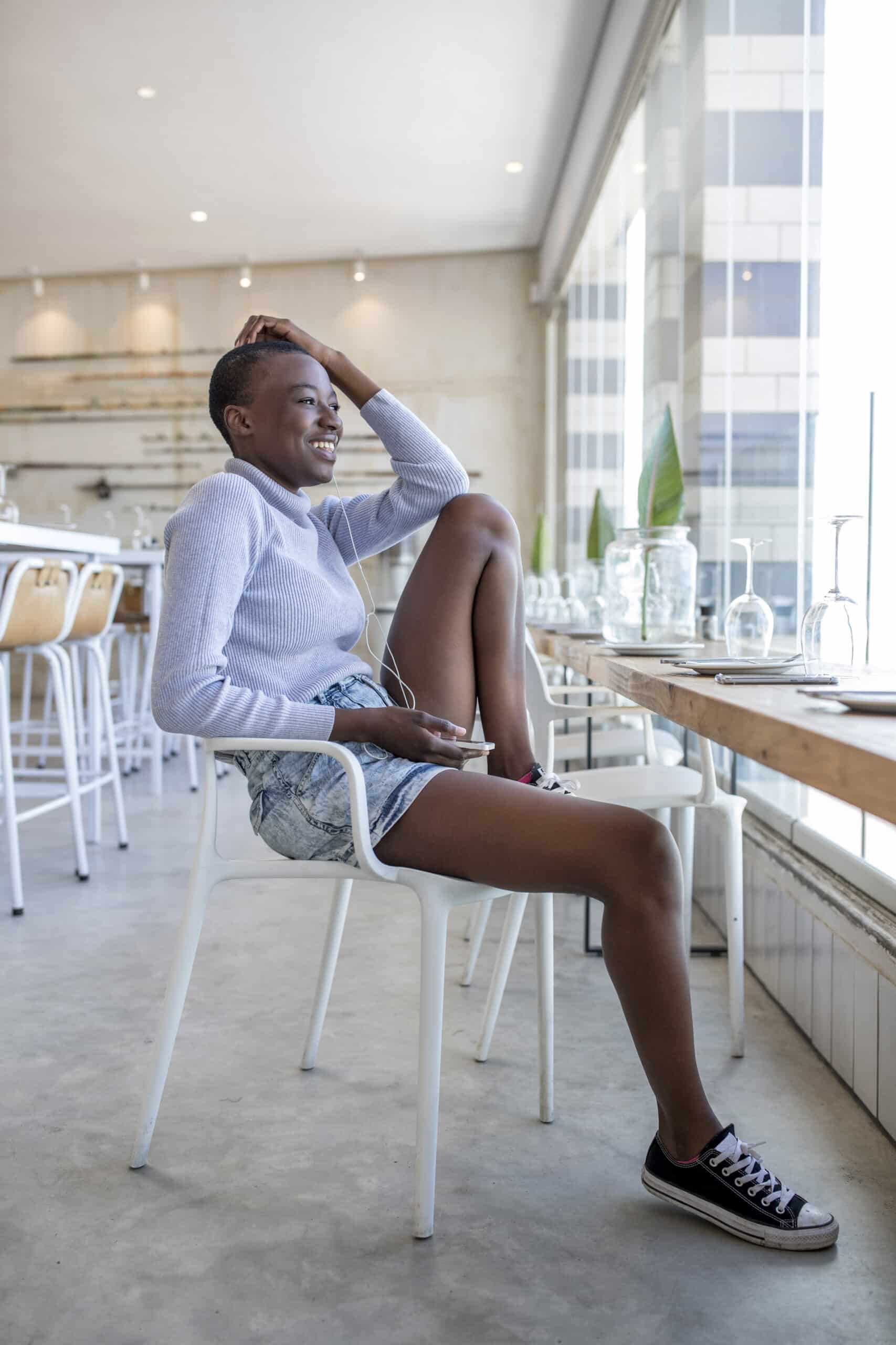 Eine fröhliche junge Frau sitzt an der hell erleuchteten Theke eines Cafés. Sie trägt einen weißen Pullover, Jeansshorts und schwarze Turnschuhe, ihre Hand ruht auf ihrem Kopf und sie lächelt. © Fotografie Tomas Rodriguez