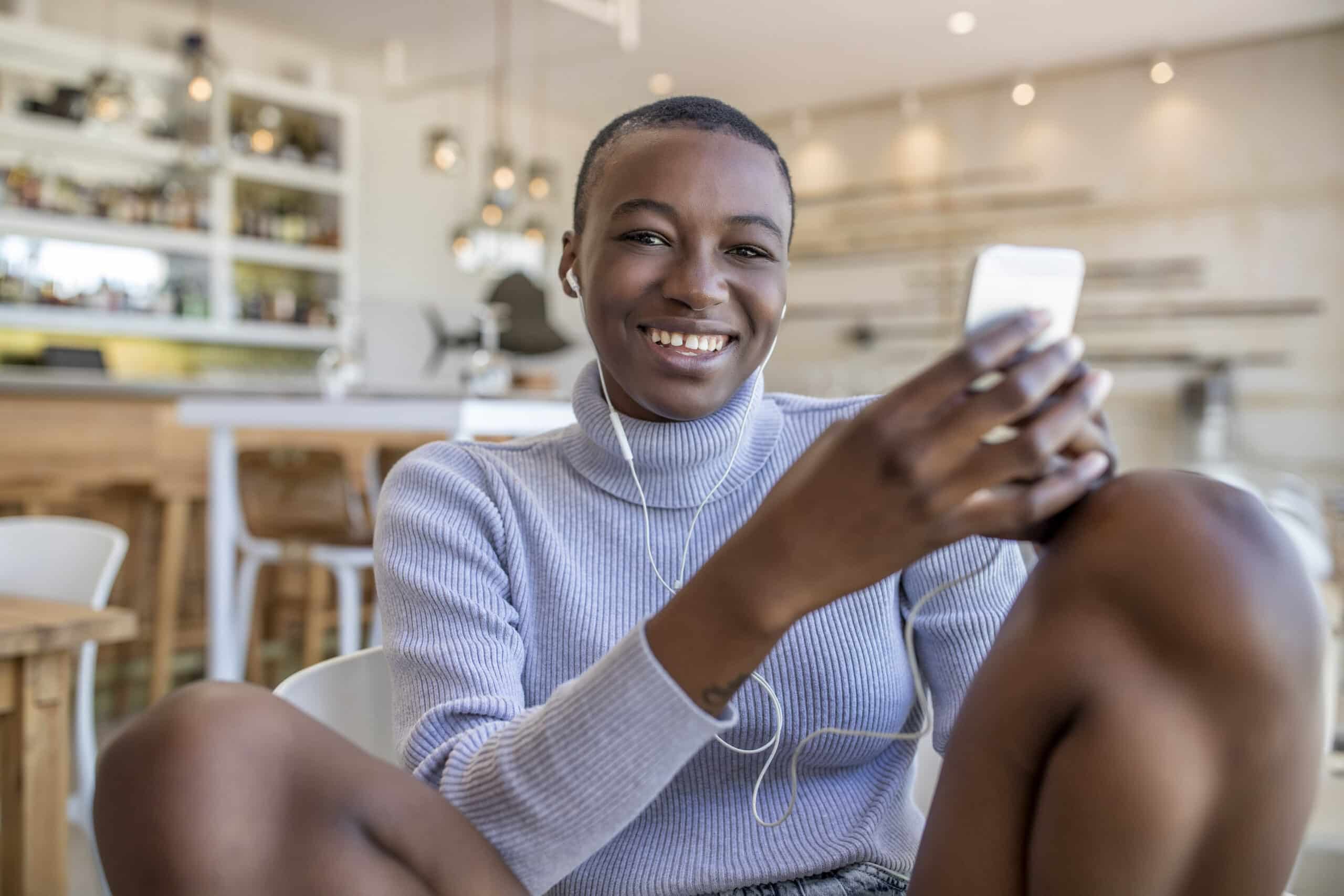 Eine fröhliche junge Frau sitzt in einem Café mit modernem, hellem Interieur und benutzt ein Smartphone. Sie lächelt in die Kamera, trägt einen lässigen Pullover und strahlt eine entspannte und glückliche Ausstrahlung aus. © Fotografie Tomas Rodriguez