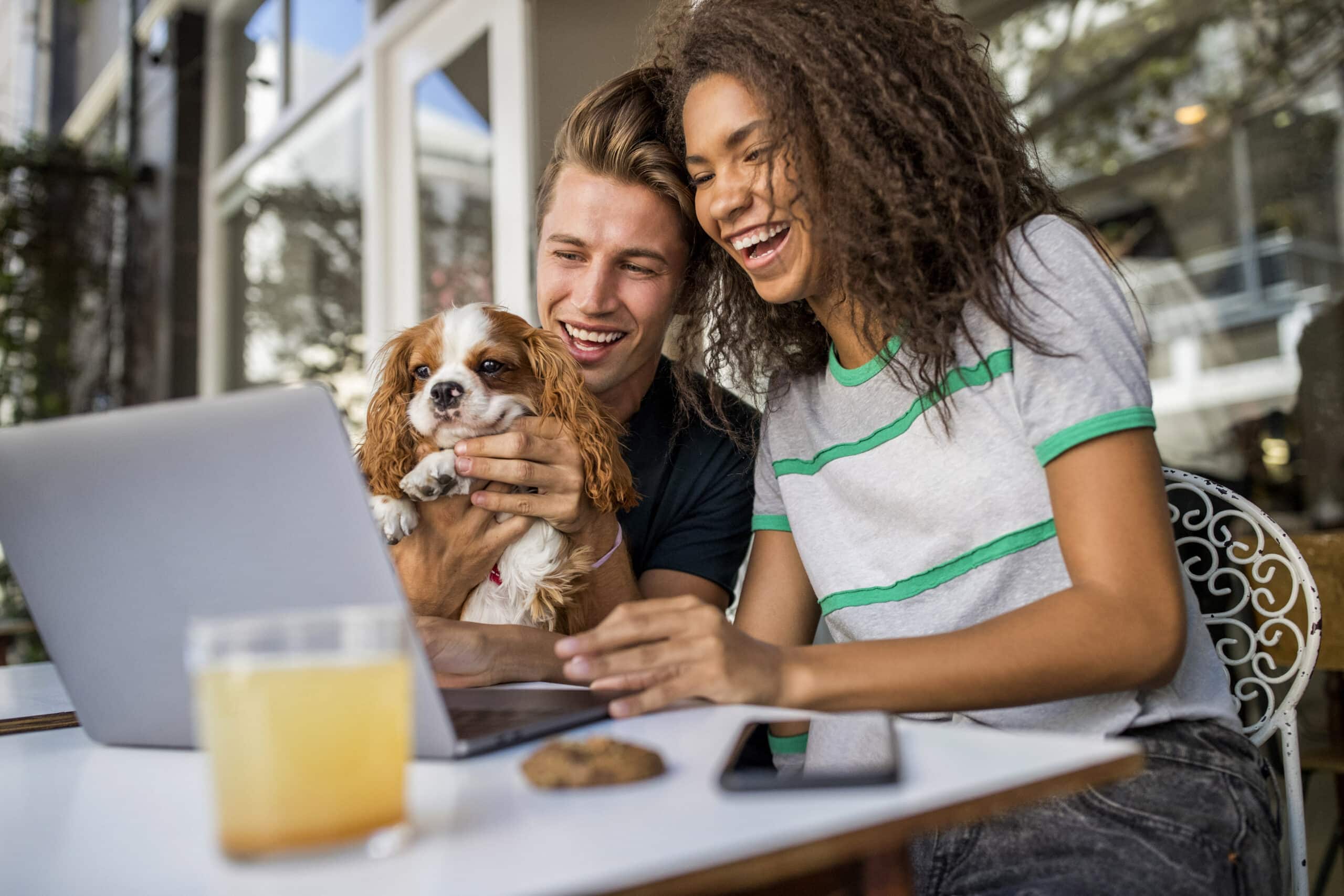 Ein fröhliches junges Paar mit einem kleinen Hund unterhält sich an einem Tisch im Freien in einem Café über einen Laptop. In der Nähe stehen Getränke und ein Snack, bei Tageslicht. © Fotografie Tomas Rodriguez