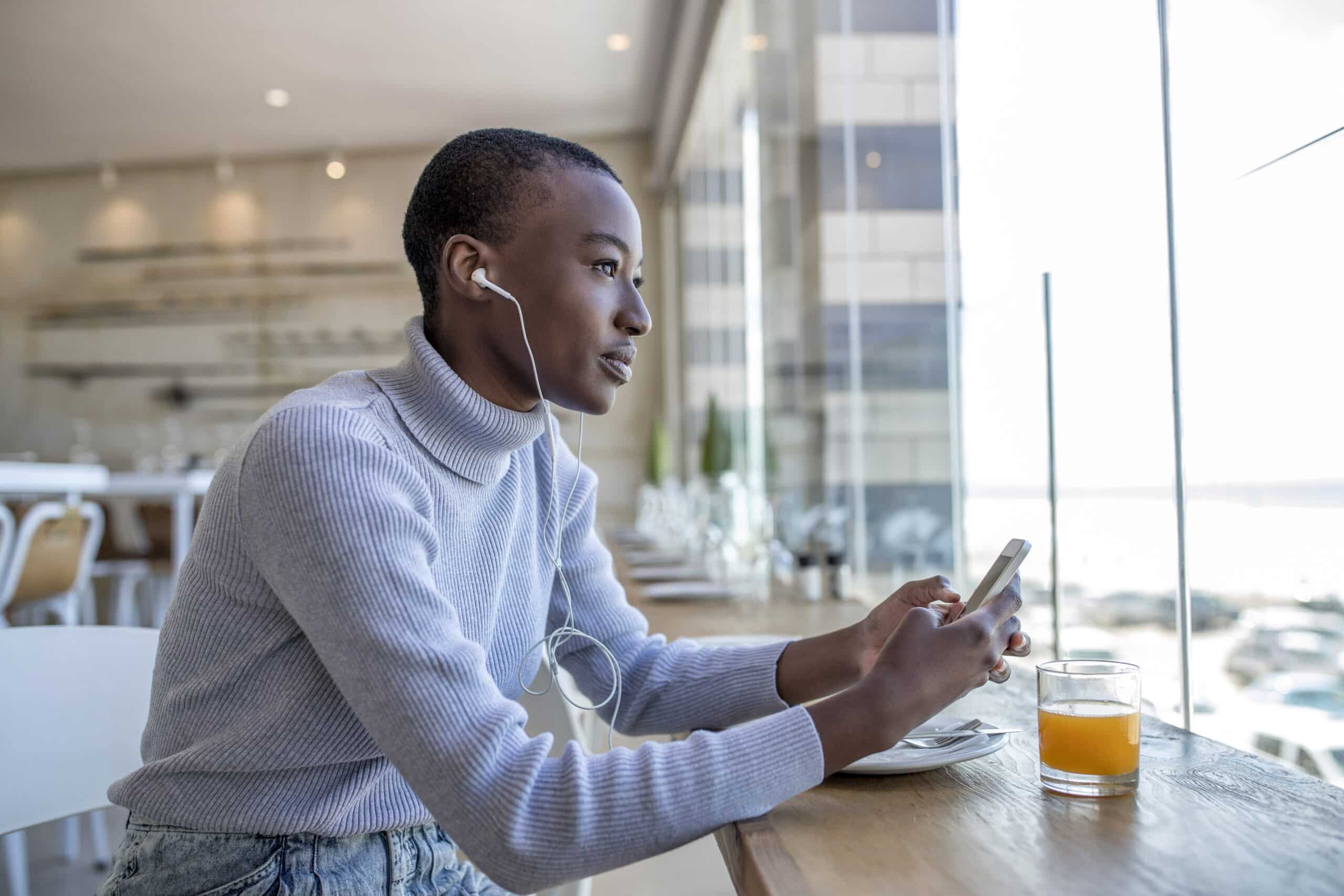 Eine junge Frau in einem hellblauen Pullover sitzt an einem Cafétisch am Fenster, benutzt ihr Smartphone und trägt Kopfhörer, neben ihr steht ein Glas Orangensaft. © Fotografie Tomas Rodriguez