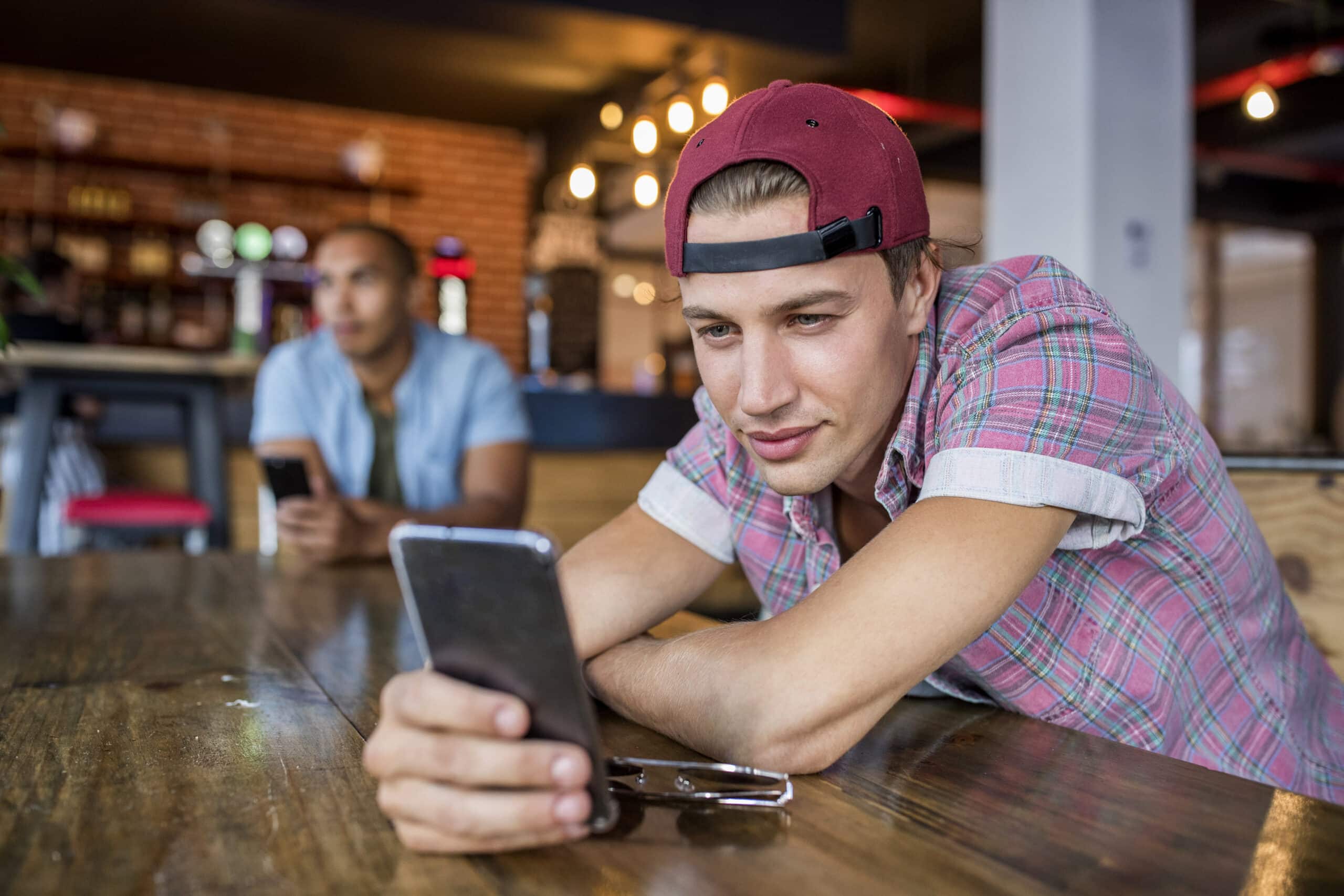 Junger Mann mit roter Mütze und kariertem Hemd benutzt sein Smartphone an einem Cafétisch, im Hintergrund ist eine andere Person unscharf zu sehen. © Fotografie Tomas Rodriguez