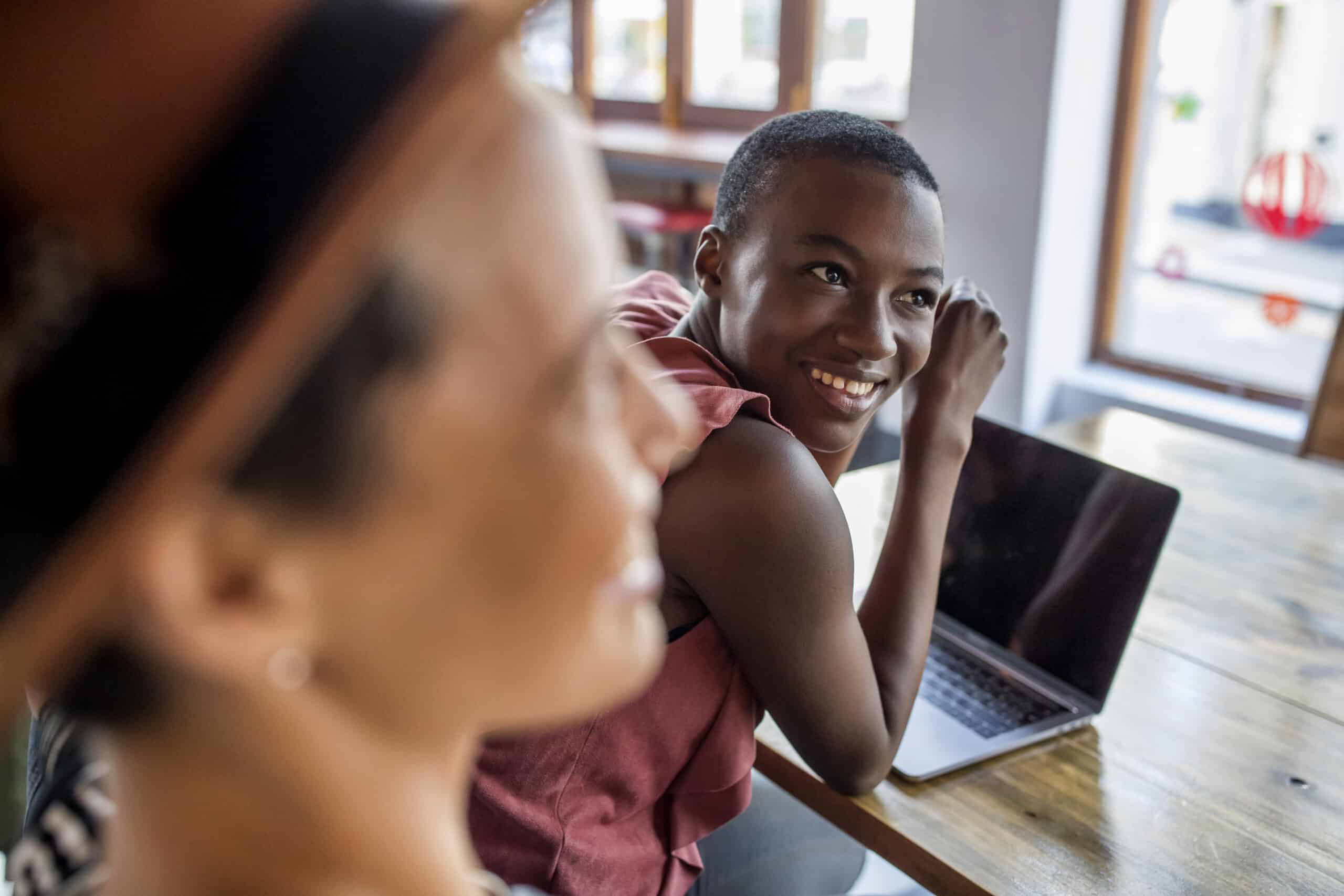 Ein junger Mann sitzt mit einem Laptop an einem Tisch und lächelt in die Kamera. Im Vordergrund ist eine unscharfe Frau zu sehen. Sie befinden sich in einer gut beleuchteten, entspannten Umgebung. © Fotografie Tomas Rodriguez
