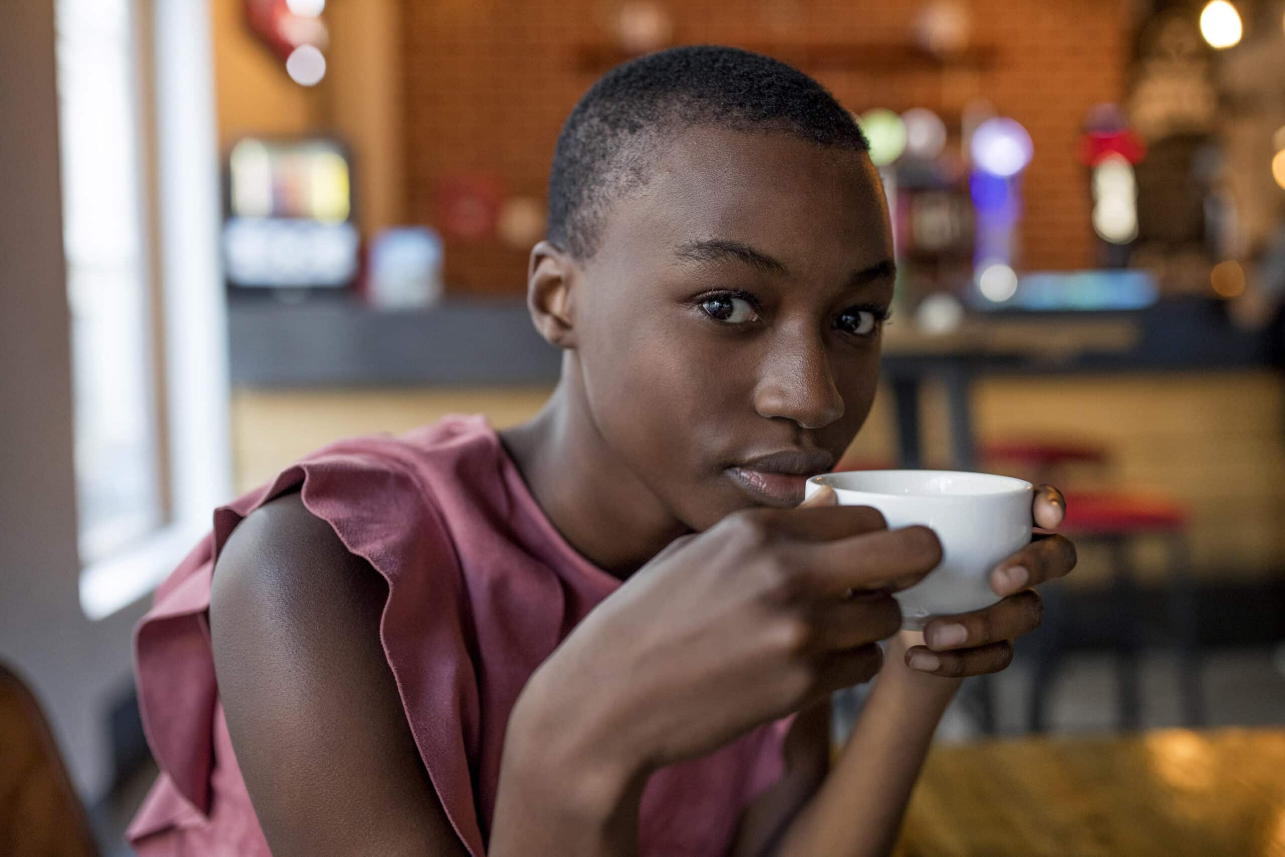 Eine junge schwarze Frau mit kurzen Haaren, einer rosa Bluse und einer weißen Tasse Kaffee in der Hand, blickt nachdenklich in die Umgebung eines Cafés. © Fotografie Tomas Rodriguez