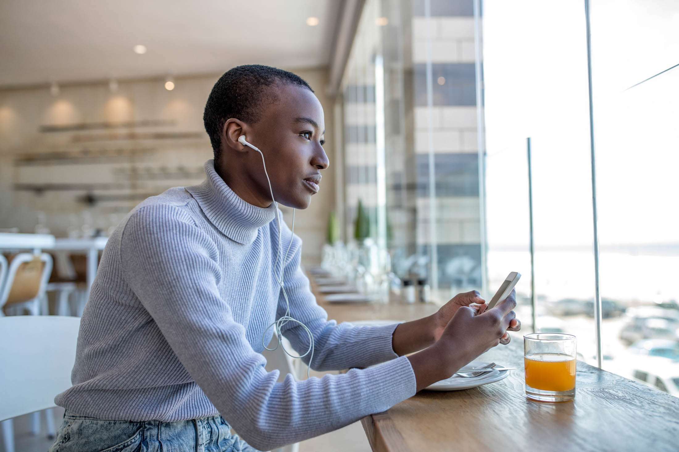 Eine junge Frau mit weißem Rollkragenpullover und Kopfhörern sitzt an einem Cafétisch am Fenster, blickt nachdenklich auf ihr Smartphone und hat neben sich ein Glas Orangensaft stehen. © Fotografie Tomas Rodriguez