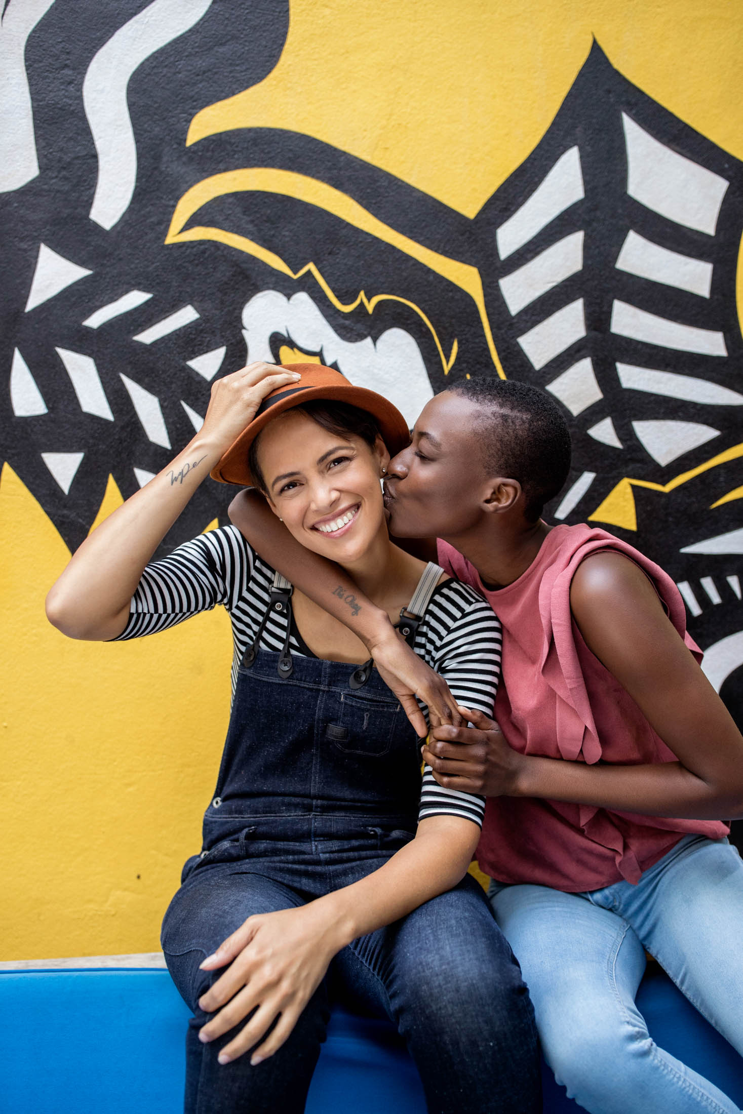 Zwei Frauen, die eine küsst die andere auf die Wange, sitzen vor einem farbenfrohen Wandgemälde. Die Frau im Overall lächelt, während sie eine Mütze zurechtrückt. Die Atmosphäre ist fröhlich und herzlich. © Fotografie Tomas Rodriguez