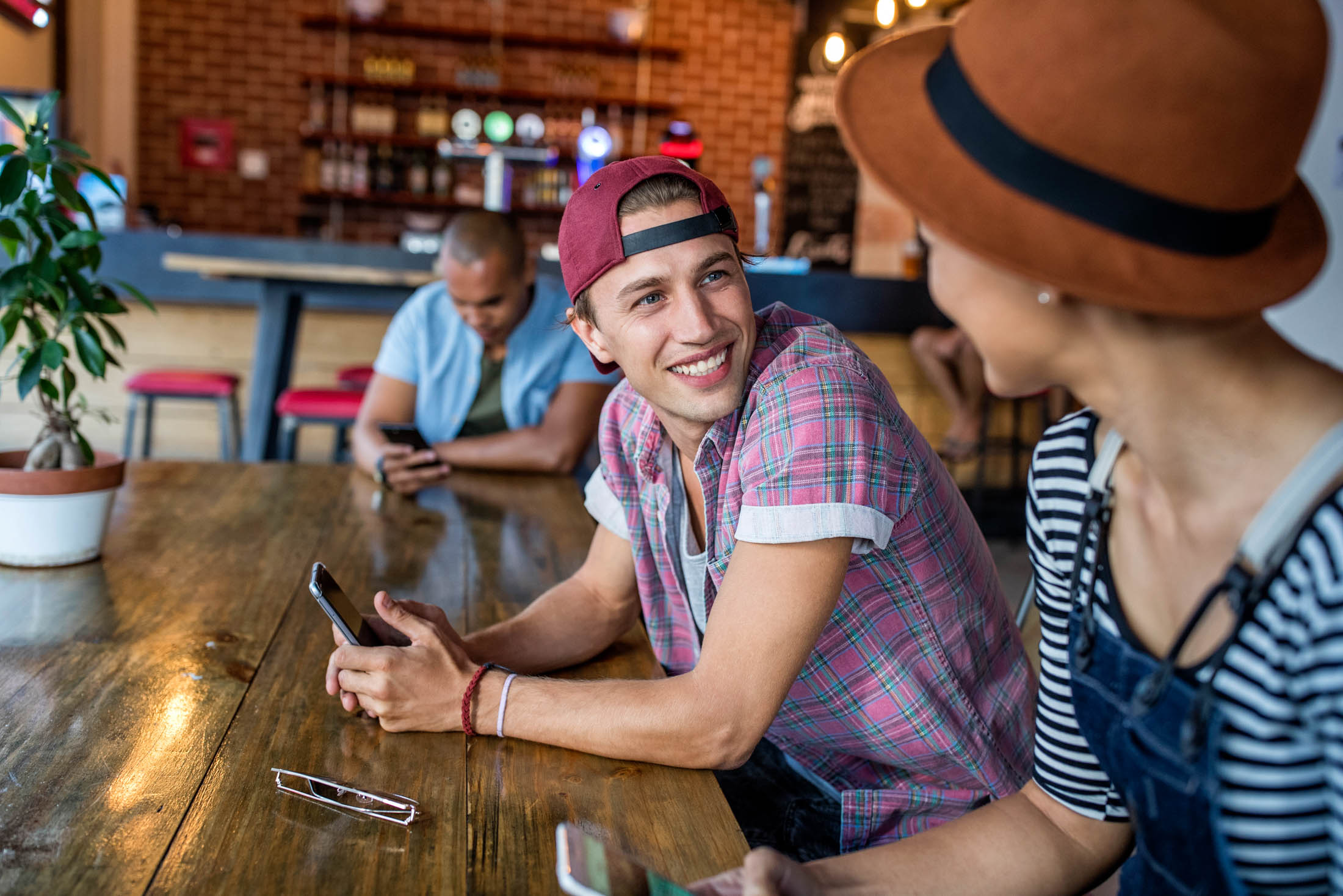 Drei junge Erwachsene sitzen in einem gemütlichen Café an einem Holztisch, plaudern und lächeln. Der junge Mann mit der Mütze hält ein Smartphone in der Hand und unterhält sich angeregt mit einer Frau neben ihm. © Fotografie Tomas Rodriguez
