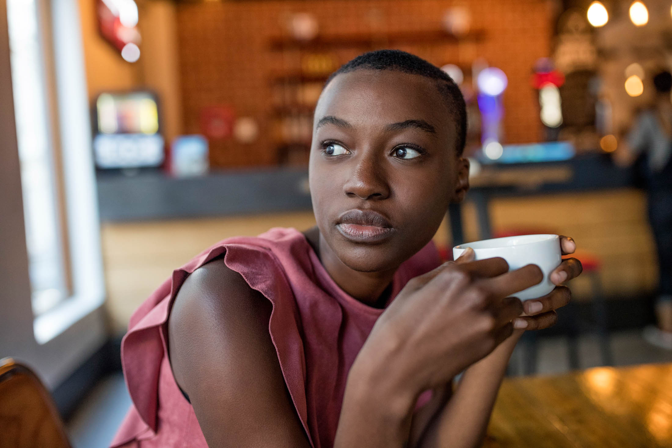 Eine nachdenkliche junge Frau hält eine Kaffeetasse und blickt in einem Café mit unscharfem Hintergrund versonnen weg. © Fotografie Tomas Rodriguez