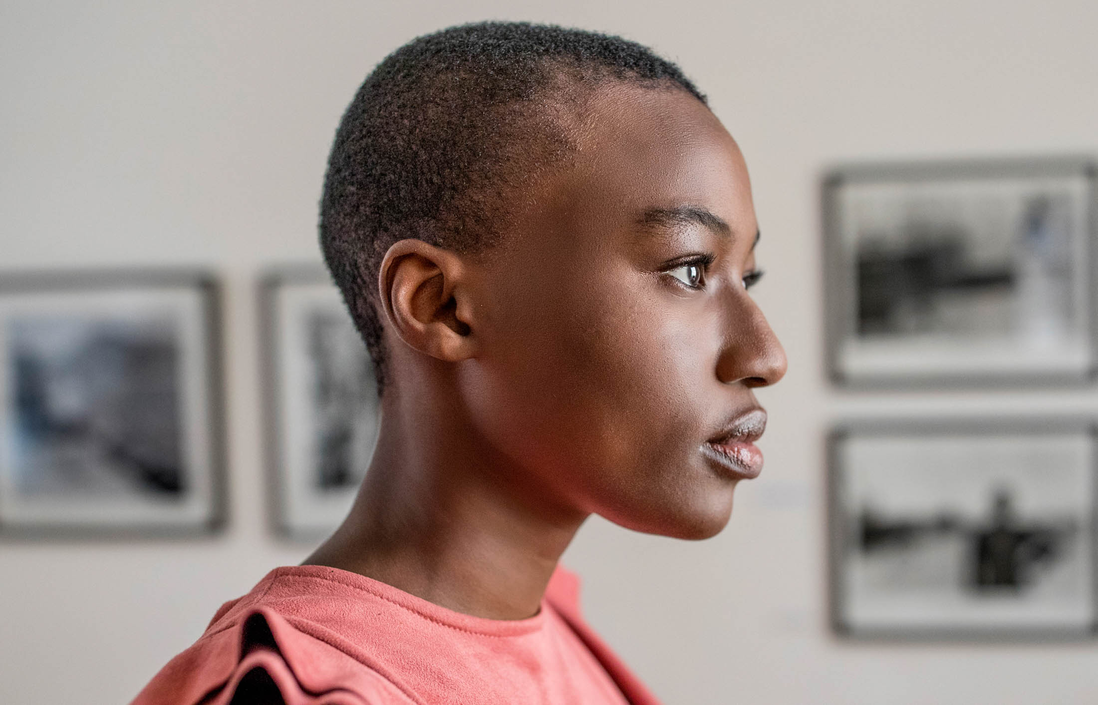 Profilansicht einer jungen Person mit kurzen Haaren und lachsfarbenem Hemd in einem Raum mit gerahmten Bildern an der Wand im Hintergrund. © Fotografie Tomas Rodriguez