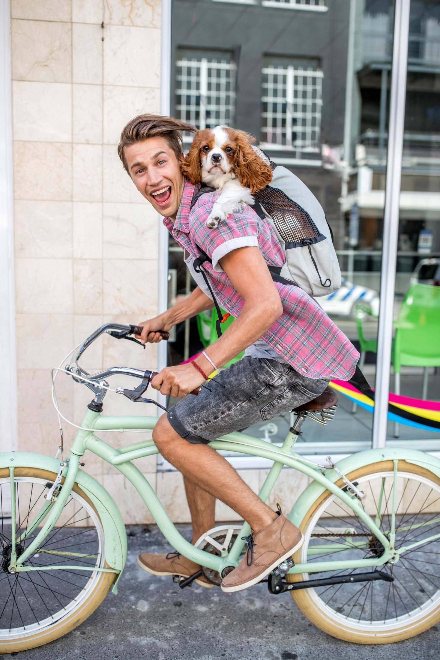 Ein fröhlicher junger Mann fährt in einer städtischen Umgebung auf einem Oldtimer-Fahrrad und hat einen kleinen Hund in einem Rucksack auf dem Rücken. Beide wirken glücklich und aufgeregt. © Fotografie Tomas Rodriguez