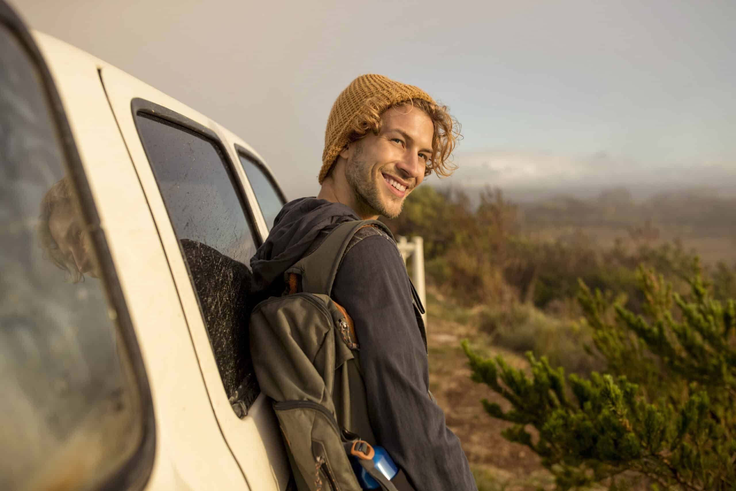 Eine Person mit lockigem Haar, einer Strickmütze und einem Rucksack lehnt lächelnd an einem weißen Lieferwagen. Sie steht an einem nebligen Tag im Freien in der Nähe einer grünen Wiese. © Fotografie Tomas Rodriguez