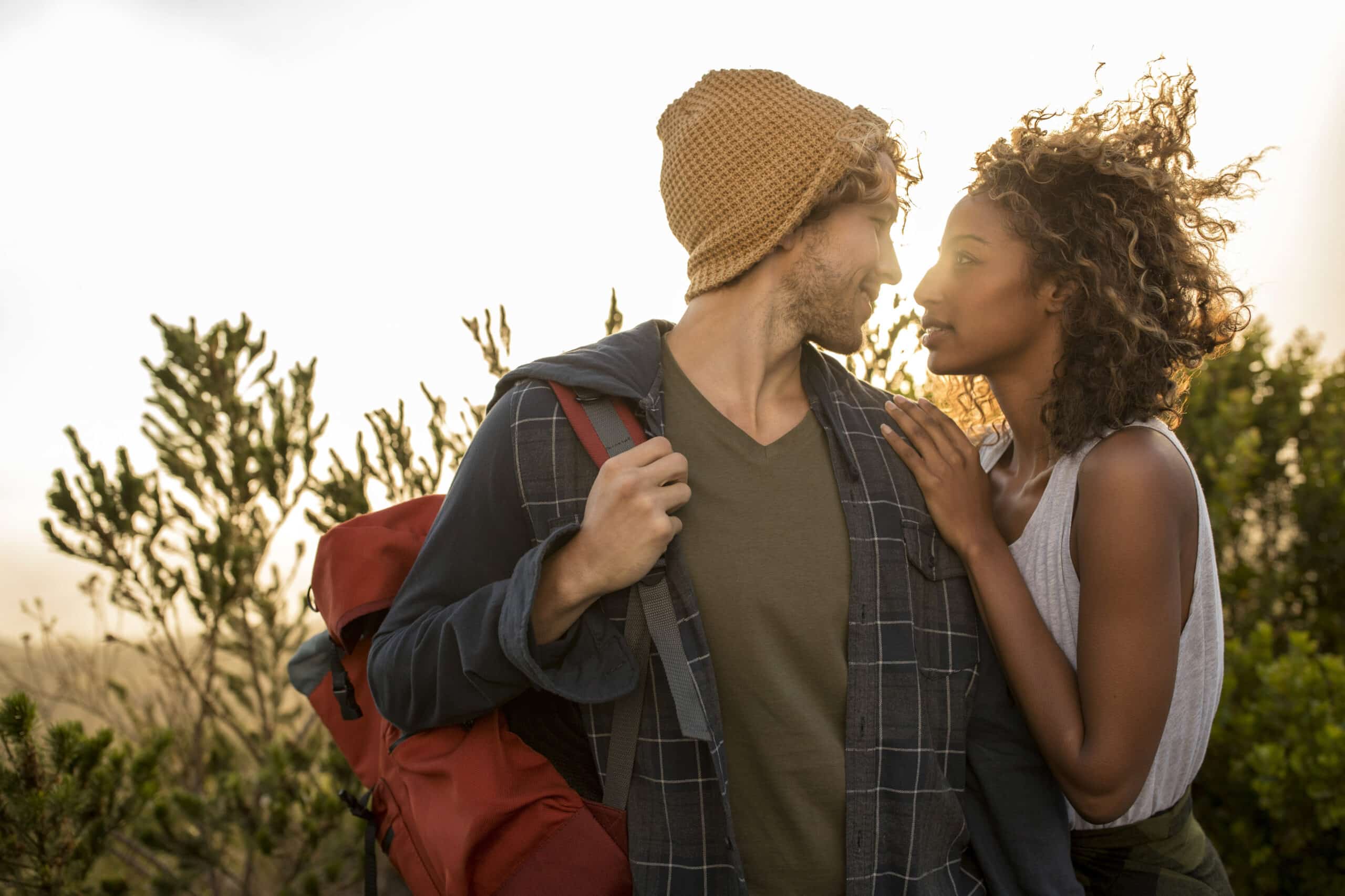 Ein Paar genießt eine Wanderung in einer Naturlandschaft. Der Mann trägt eine Beanie-Mütze und einen Rucksack, während die Frau mit dem lockigen Haar ihre Hand auf seine Schulter legt. Sie blicken einander an, während die Sonne hinter ihnen untergeht, umgeben von Grün. © Fotografie Tomas Rodriguez