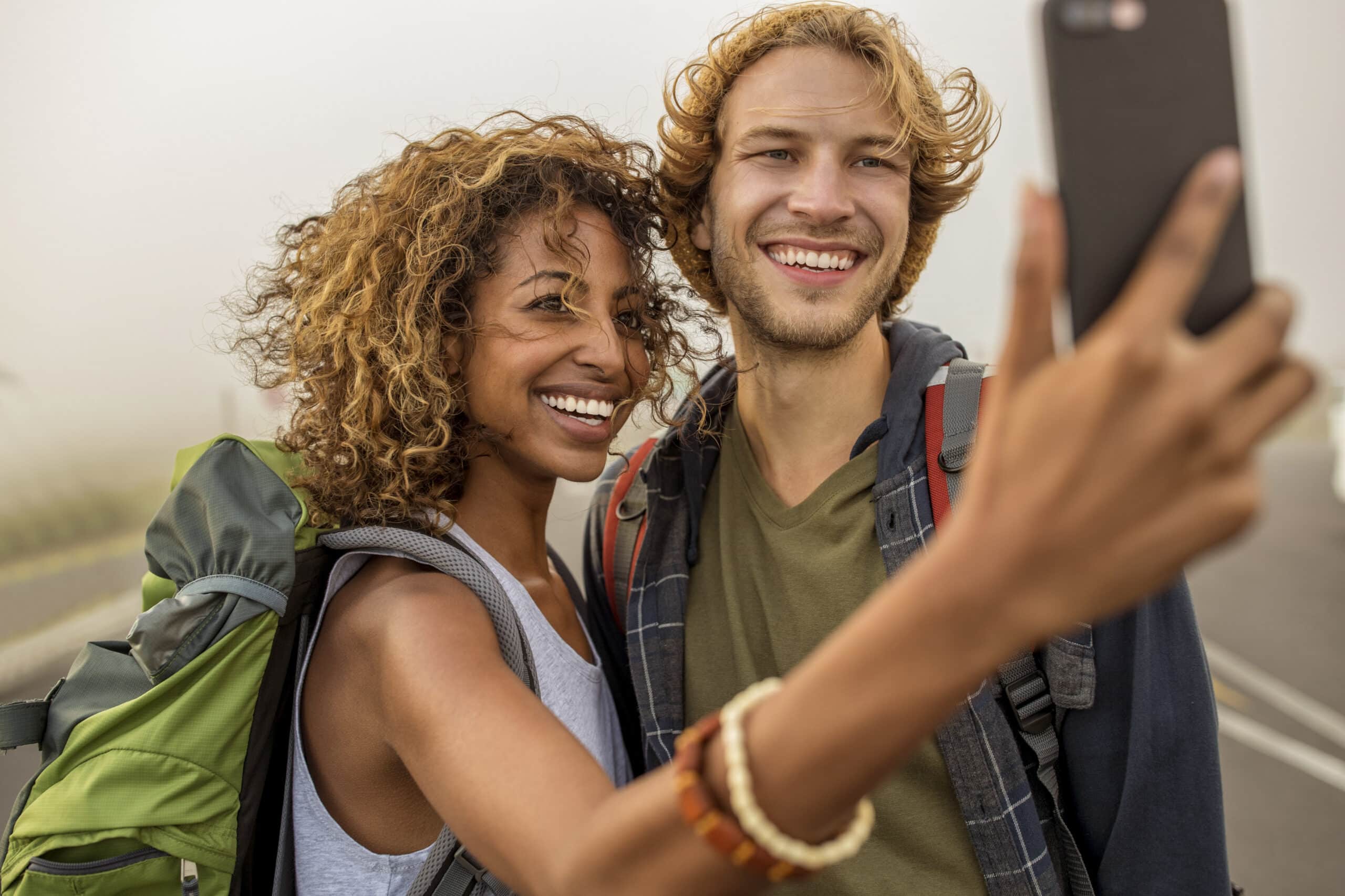 Ein junges Paar mit Rucksäcken macht ein fröhliches Selfie. Die Frau hat lockiges Haar und trägt ein grünes Oberteil, während der Mann mit welligem Haar eine Beanie und ein graues T-Shirt trägt. Sie scheinen ein Outdoor-Abenteuer an einem nebligen Tag zu genießen. © Fotografie Tomas Rodriguez