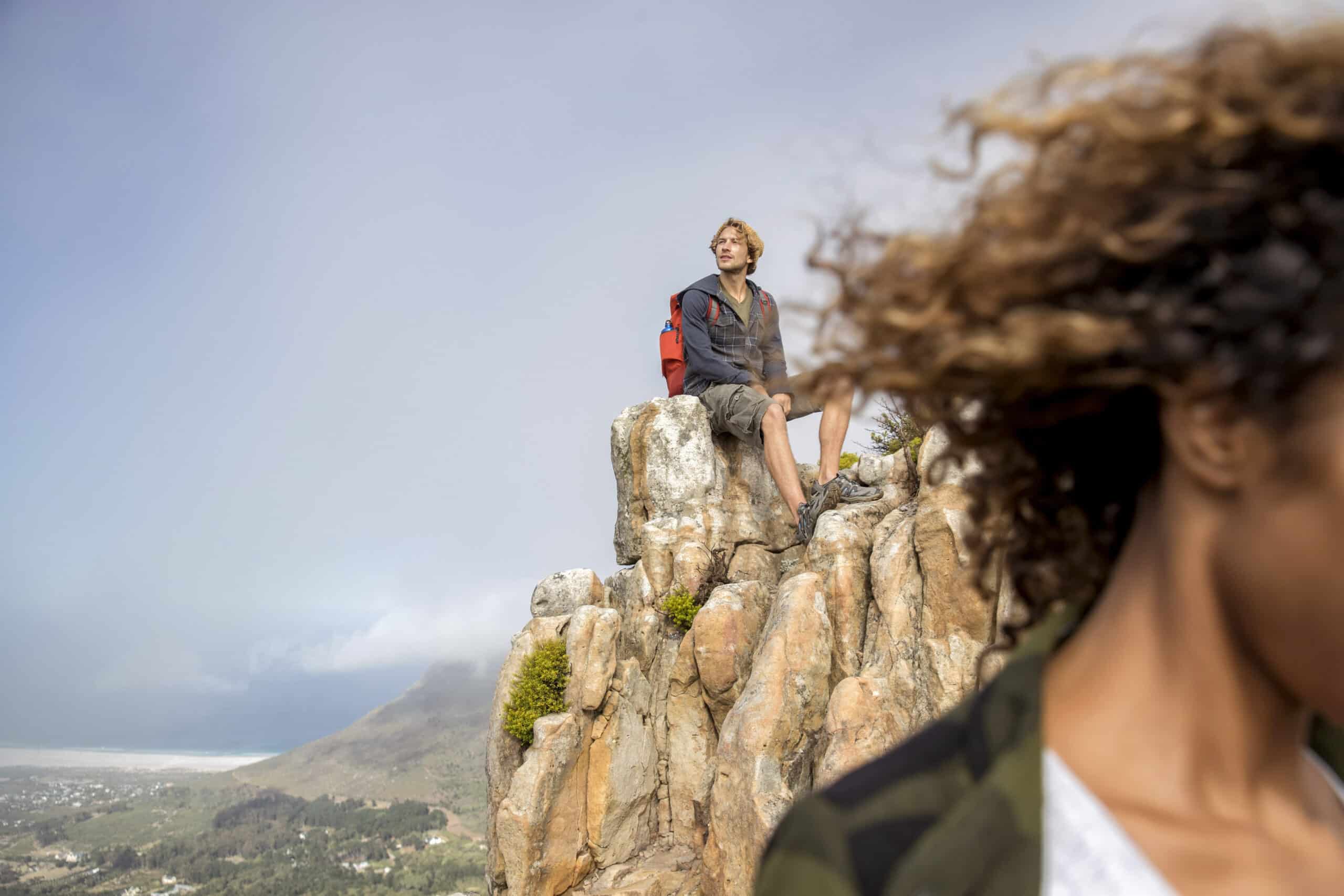 Eine Person mit Rucksack sitzt auf einem Felsvorsprung und blickt nach oben. Eine weitere Person mit lockigem Haar und Jacke ist im Vordergrund teilweise sichtbar, ihr Gesicht ist von der Kamera abgewandt. Im Hintergrund sind Hügel und ein bewölkter Himmel zu sehen. © Fotografie Tomas Rodriguez