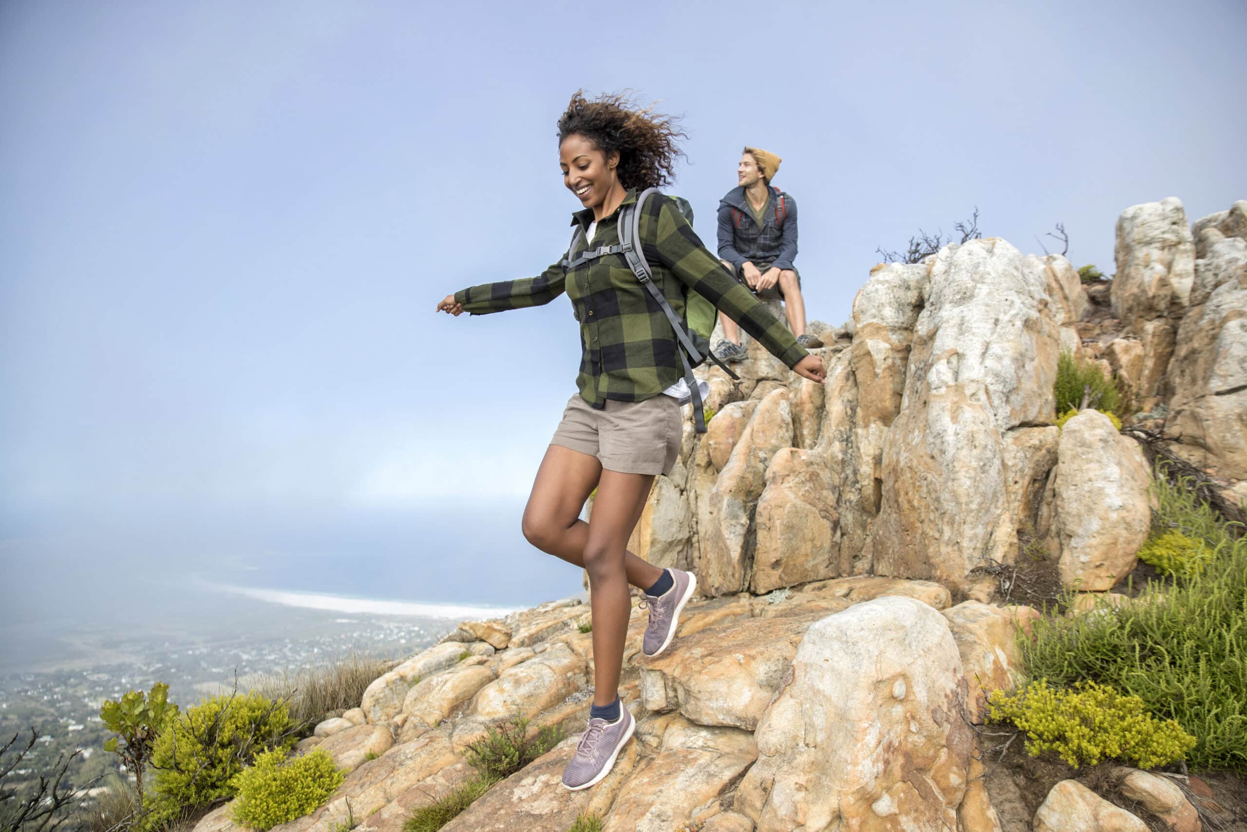 Eine Frau springt freudig von einem Felsen zum anderen auf einer Felsklippe, während eine andere Person hinter ihr sitzt und die Aussicht genießt. Die Landschaft darunter umfasst entfernte Vegetation und einen dunstigen Himmel. Beide Personen tragen legere Outdoor-Kleidung. © Fotografie Tomas Rodriguez
