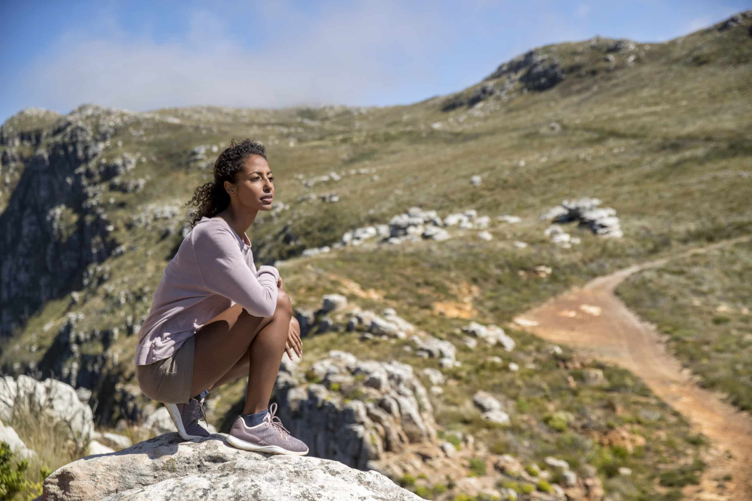 Eine Person mit lockigem Haar hockt mit geschlossenen Augen auf einem Felsen und trägt einen rosa-beigen Pullover und Shorts mit passenden Turnschuhen. Sie befinden sich in einer malerischen Berggegend mit felsigem Gelände und einem klaren Pfad im Hintergrund unter einem teilweise bewölkten Himmel. © Fotografie Tomas Rodriguez