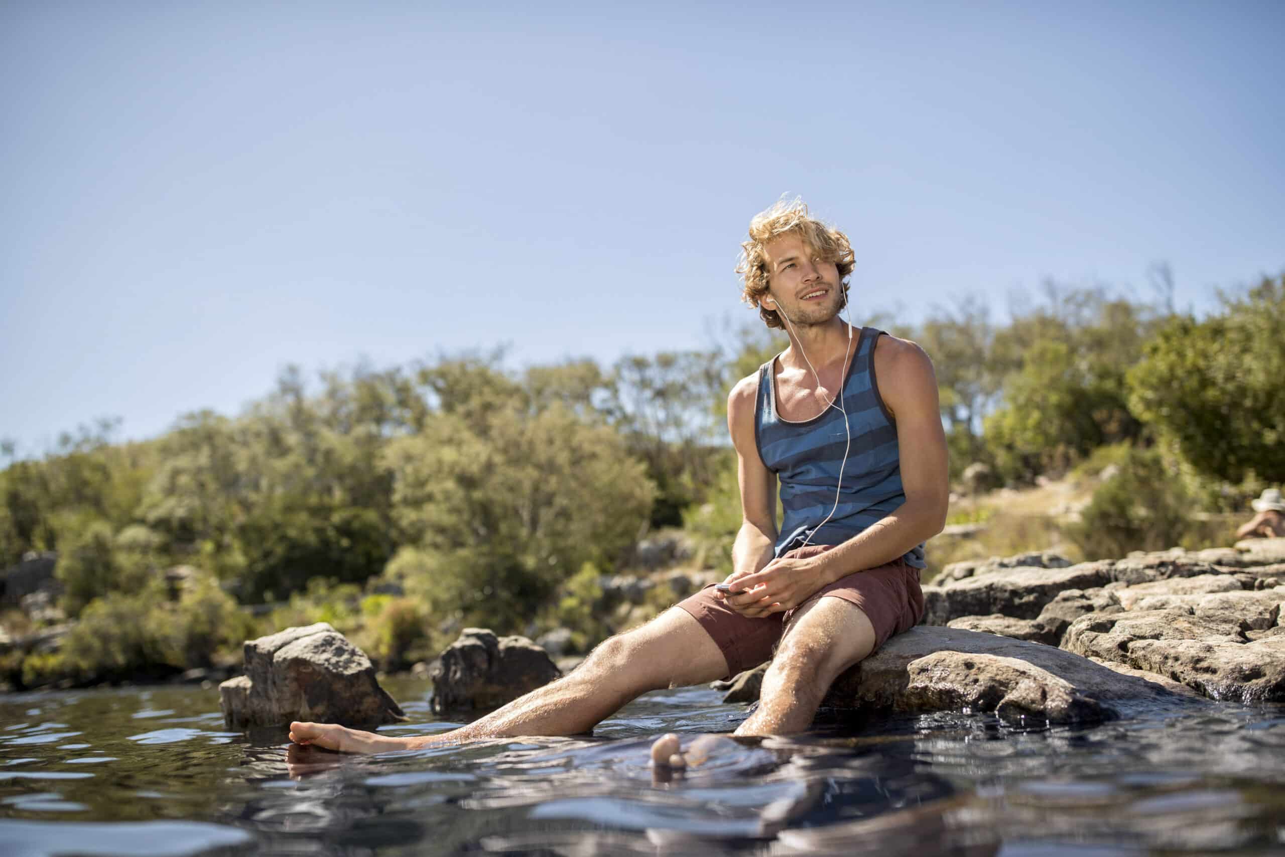 Ein Mann mit blondem Haar, der ein blaues Tanktop und braune Shorts trägt, sitzt auf einem Felsen am Wasser, seine Füße sind im Wasser. Er hört über Kopfhörer Musik und blickt in die Ferne, umgeben von Grün unter einem klaren blauen Himmel. © Fotografie Tomas Rodriguez
