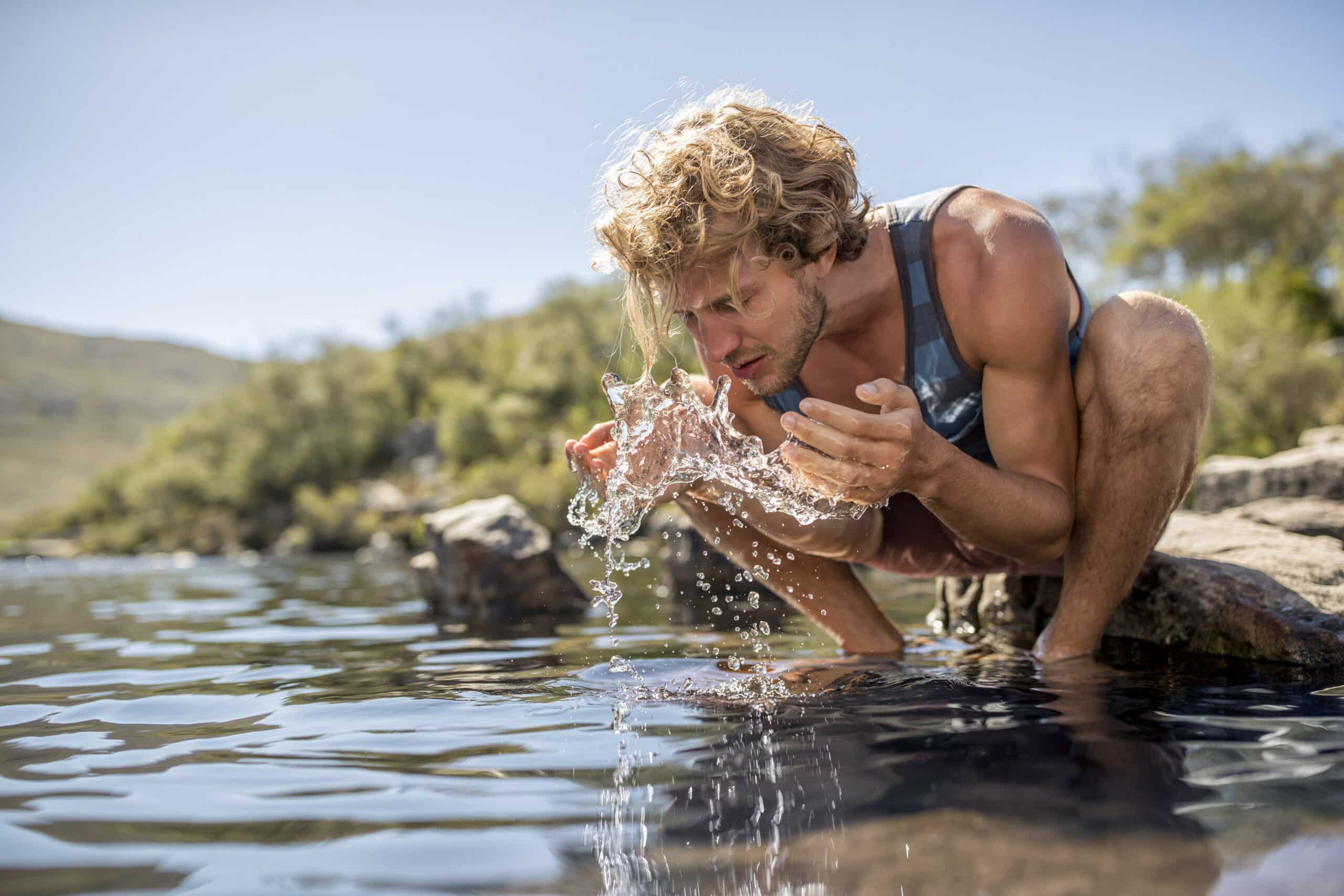 Ein Mann mit blondem Haar kniet neben einem klaren Bach, schöpft Wasser in seine Hände und spritzt sich damit ins Gesicht. Er trägt ein ärmelloses Hemd und Shorts, vor der Kulisse grüner Blätter und entfernter Hügel unter einem klaren blauen Himmel. © Fotografie Tomas Rodriguez