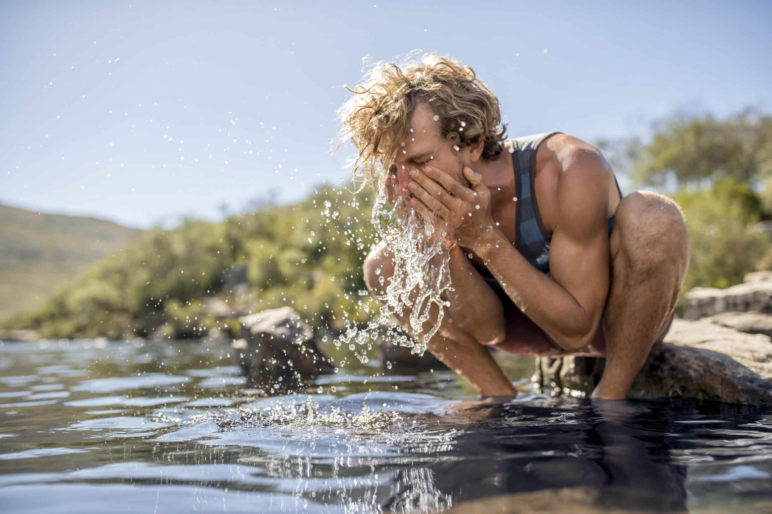 Eine Person mit blonden Haaren und einem Tanktop hockt neben einem Fluss und spritzt sich mit den Händen Wasser ins Gesicht. Die Szene spielt im Freien mit Felsen, Grün und einem klaren Himmel im Hintergrund. © Fotografie Tomas Rodriguez