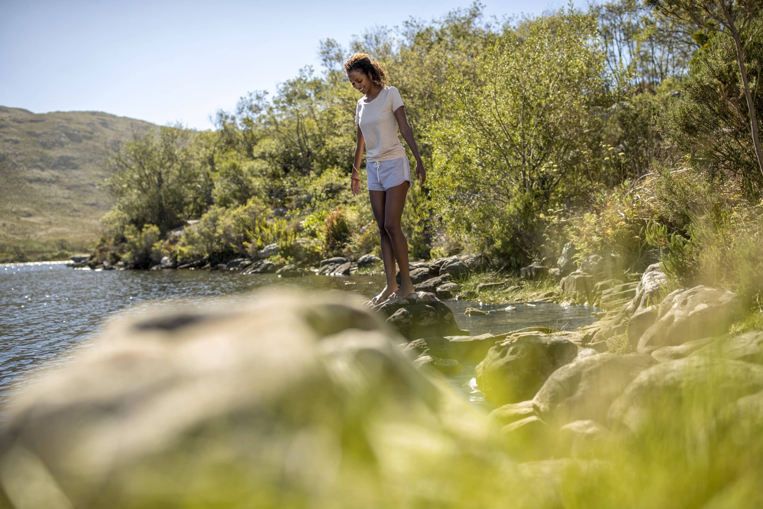 Eine Frau in weißem Hemd und Shorts steht auf Felsen an einem ruhigen, von Grün umgebenen See. Sie blickt auf das Wasser hinunter, im Hintergrund sind Berge unter einem klaren blauen Himmel zu sehen. © Fotografie Tomas Rodriguez