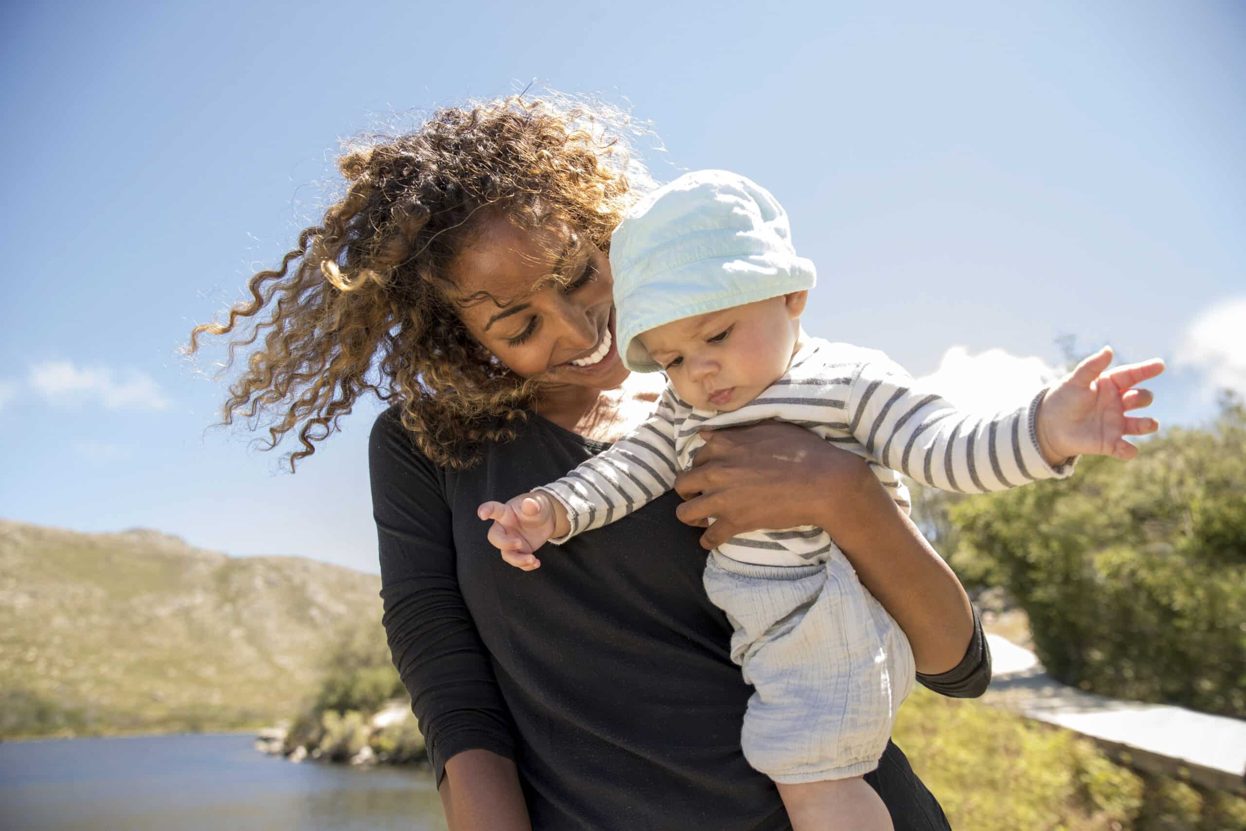 Eine Frau mit lockigem Haar und einem schwarzen Langarmshirt lächelt ein Baby in ihren Armen an. Das Baby, das eine hellblaue Mütze und ein gestreiftes Shirt trägt, schaut mit ausgestreckten Armen nach unten. Sie sind an einem sonnigen Tag im Freien mit einer malerischen Kulisse aus Hügeln, Wasser und einem klaren blauen Himmel. © Fotografie Tomas Rodriguez