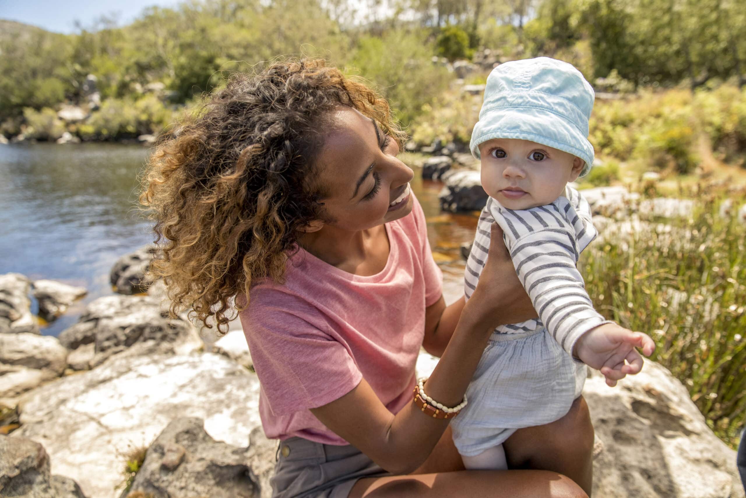 Eine Frau mit lockigem Haar und einem rosa Hemd lächelt ein Baby an, das eine blaue Mütze und ein gestreiftes Hemd trägt. Sie sind draußen in der Nähe eines Gewässers, umgeben von Felsen und Grün. © Fotografie Tomas Rodriguez