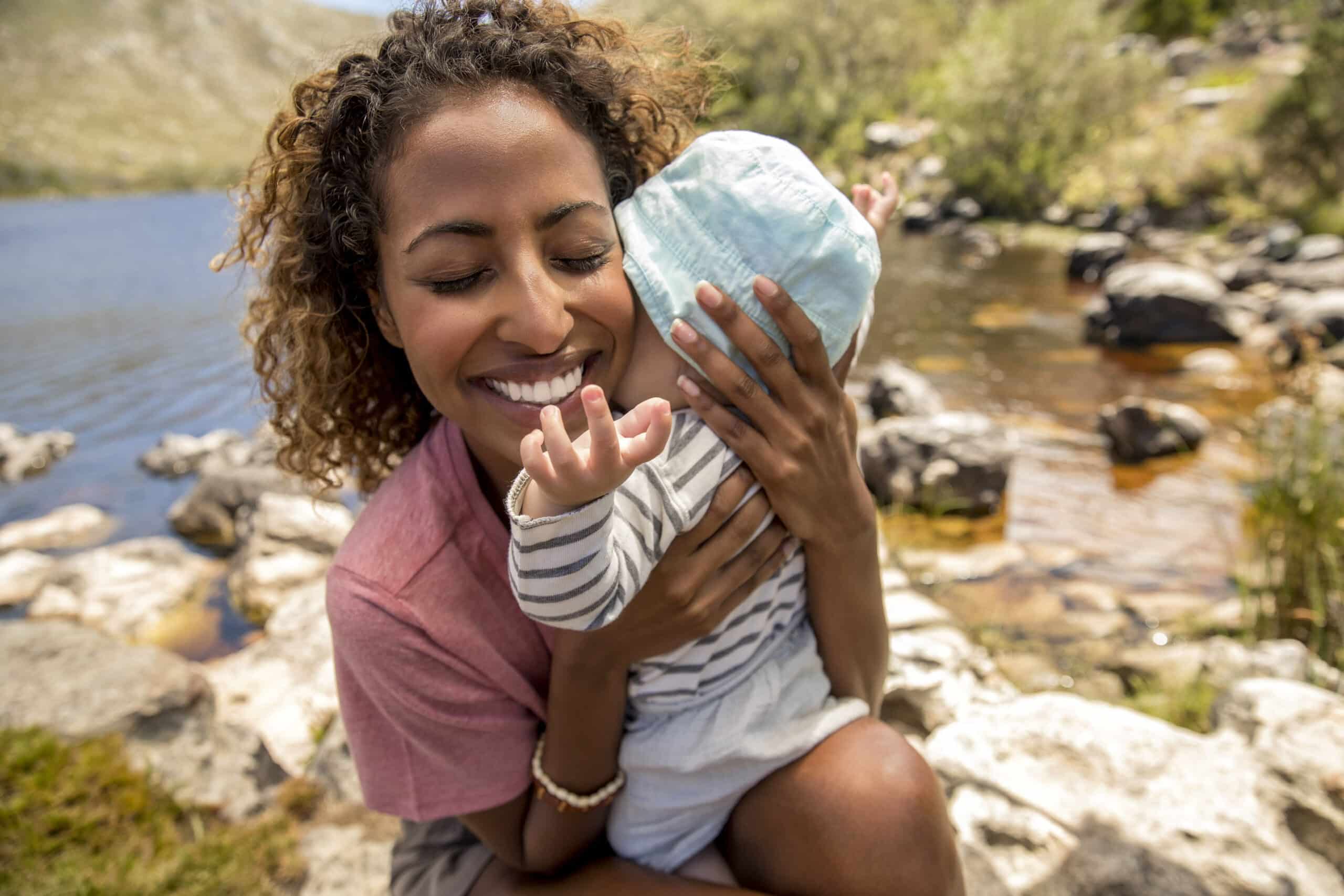 Eine Frau mit lockigem Haar und einem breiten Lächeln hält ein Baby mit Sonnenhut in der Nähe eines Sees mit felsigem Ufer. Das Baby streckt sich nach oben, um das Gesicht der Frau zu berühren. Im Hintergrund sind Wasser und Grün unter einem sonnigen Himmel zu sehen. © Fotografie Tomas Rodriguez
