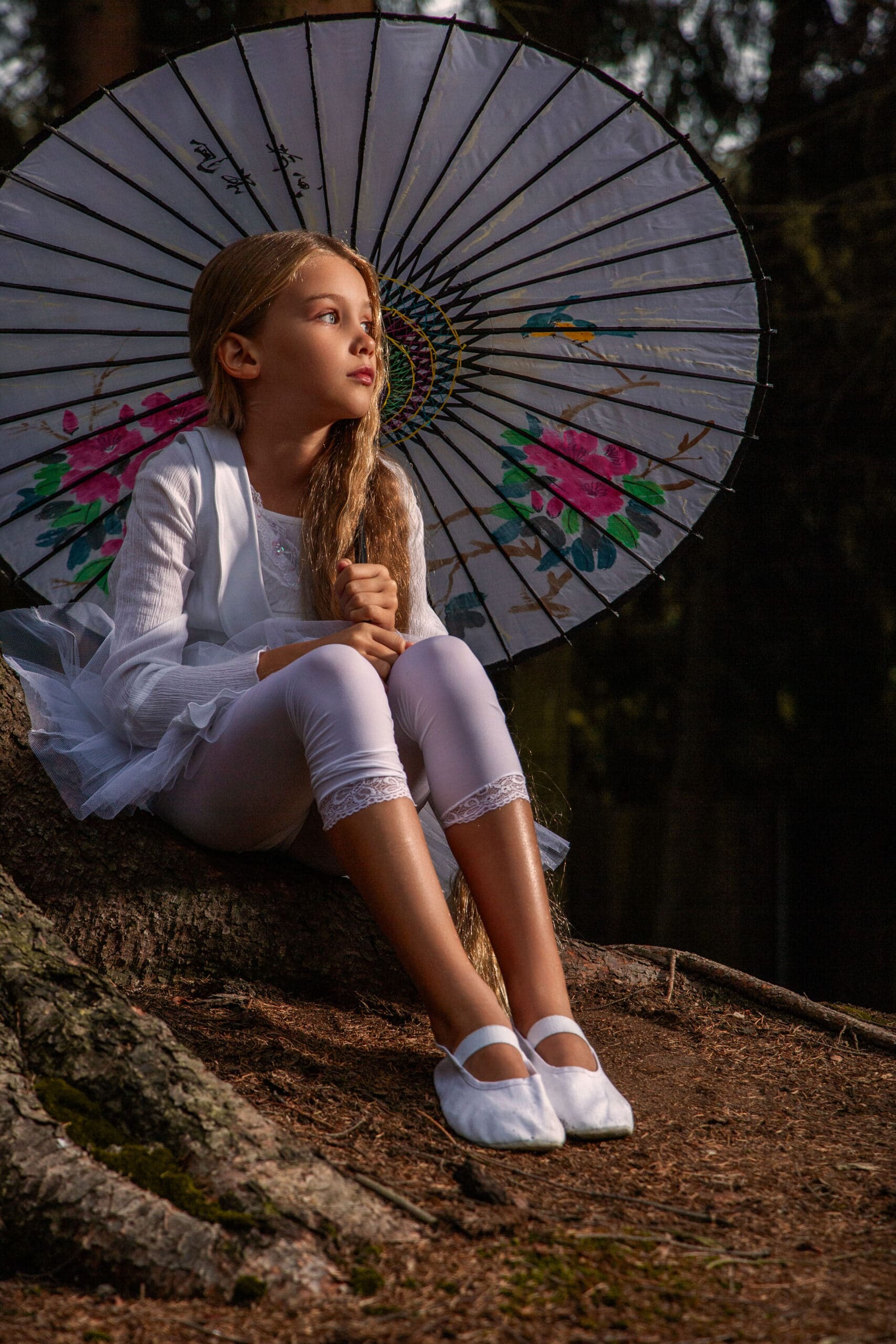 Ein junges Mädchen sitzt unter einem Baum und hält einen bunten Fächer hinter sich. Sie trägt ein weißes Kleid und blickt nachdenklich in die Ferne. Die Umgebung ist heiter, gedämpftes Tageslicht hebt sie hervor. © Fotografie Tomas Rodriguez