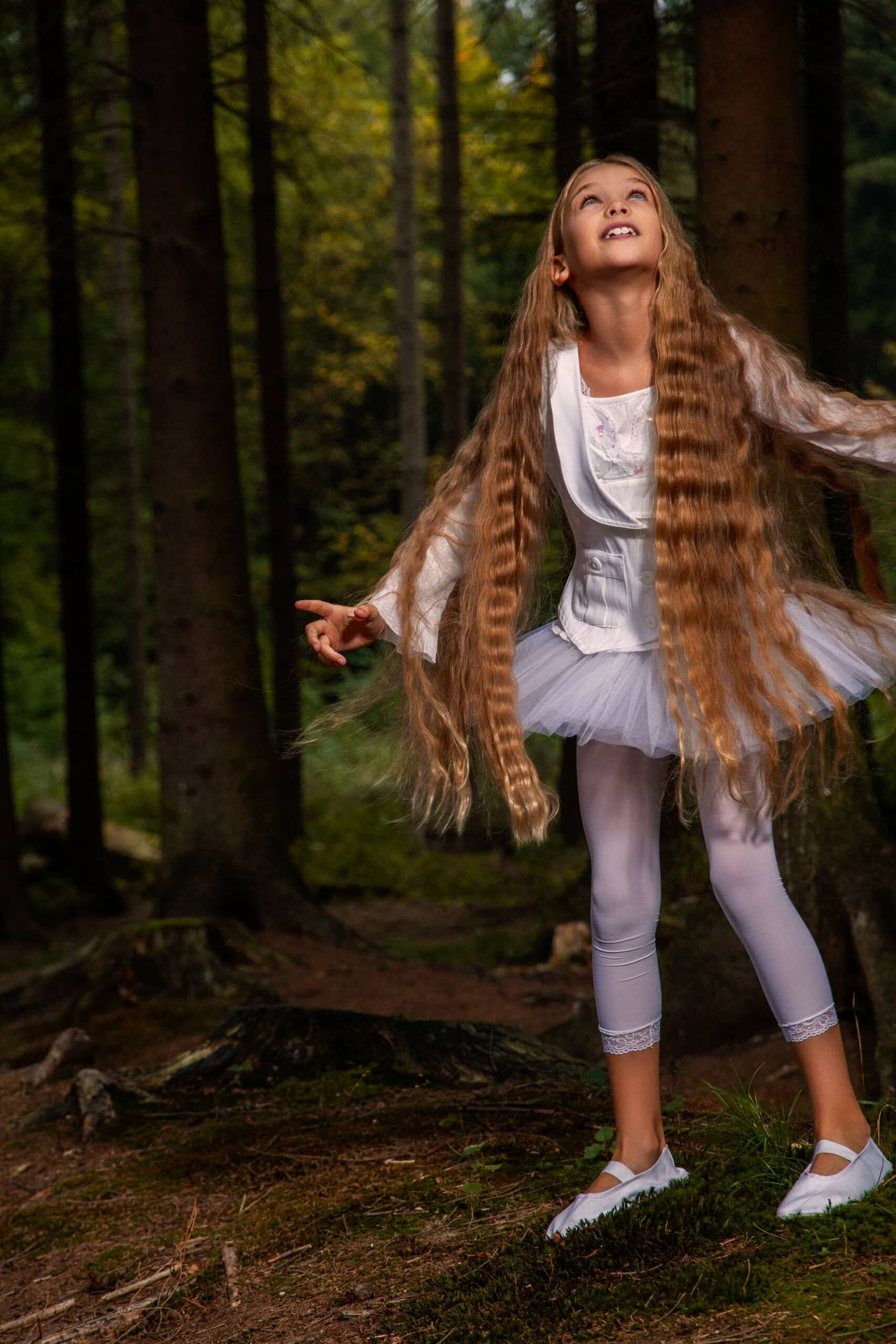 Ein junges Mädchen mit langem, wallendem Haar, einem weißen Kleid und Leggings, blickt mit freudiger Miene zwischen hohen, schattigen Bäumen in einem Wald nach oben. © Fotografie Tomas Rodriguez