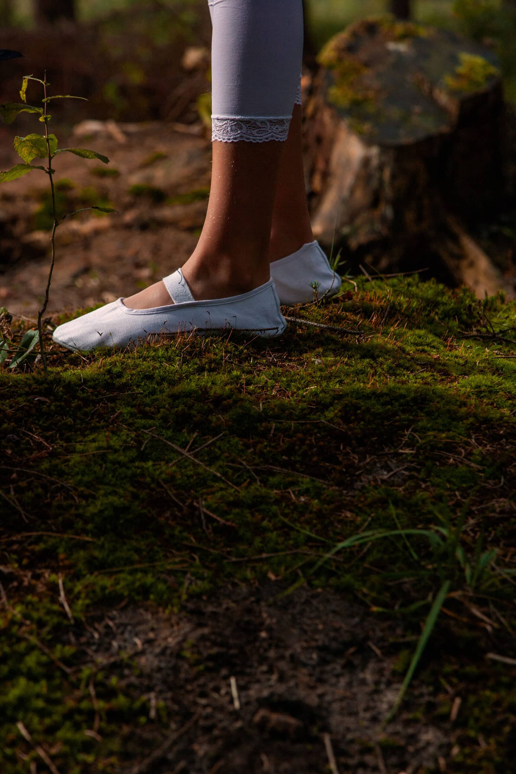 Nahaufnahme der Füße einer Person in weißen Slippern und Socken mit Spitzenrand. Sie steht auf einem moosigen Waldboden. In der Nähe steht ein Baumstumpf, beleuchtet durch warmes Sonnenlicht. © Fotografie Tomas Rodriguez