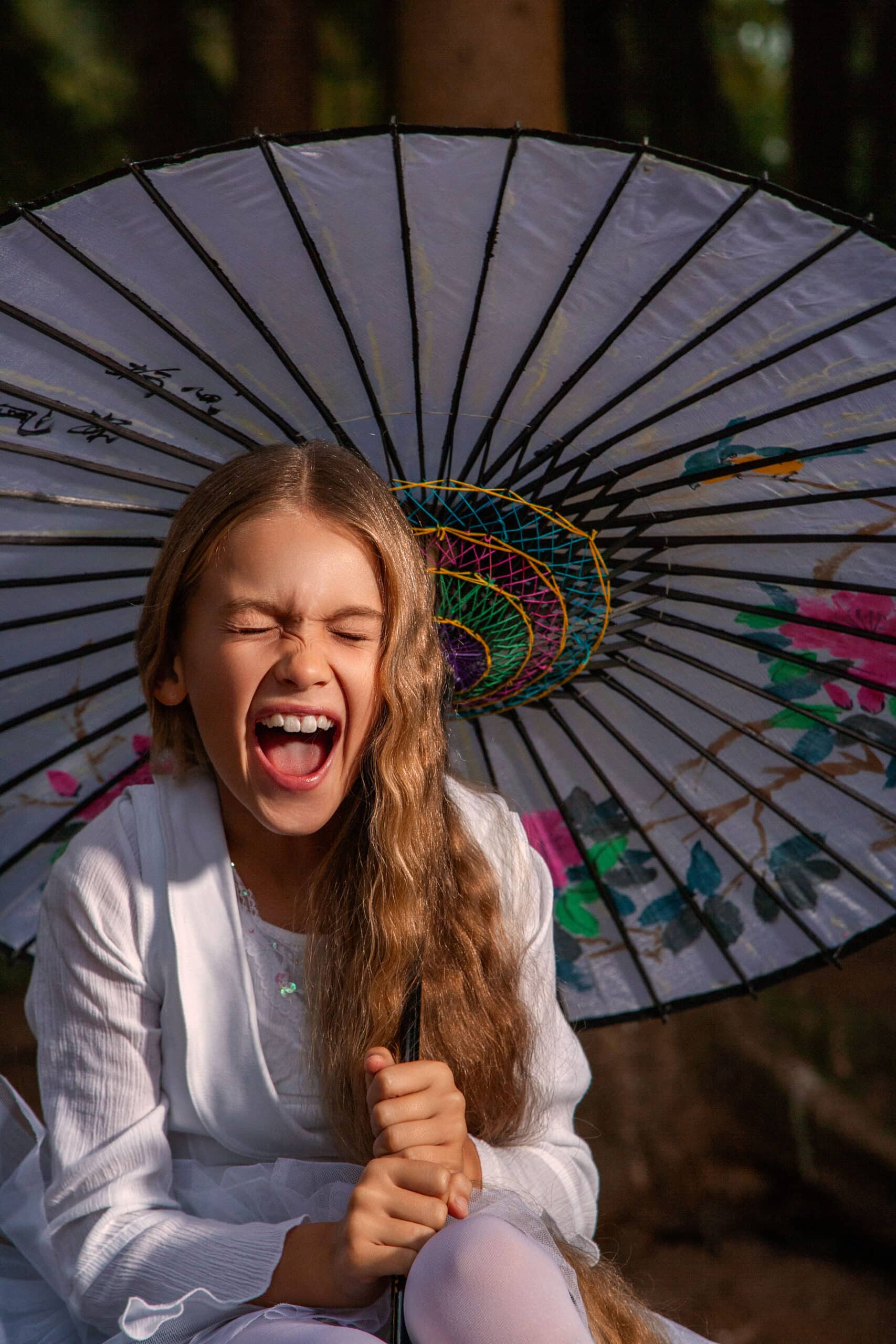 Ein junges Mädchen mit langen braunen Haaren lacht fröhlich, hält einen dekorativen weißen Regenschirm mit schwarzen und regenbogenfarbenen Akzenten und sitzt in einer sonnenbeschienenen Waldlandschaft. © Fotografie Tomas Rodriguez