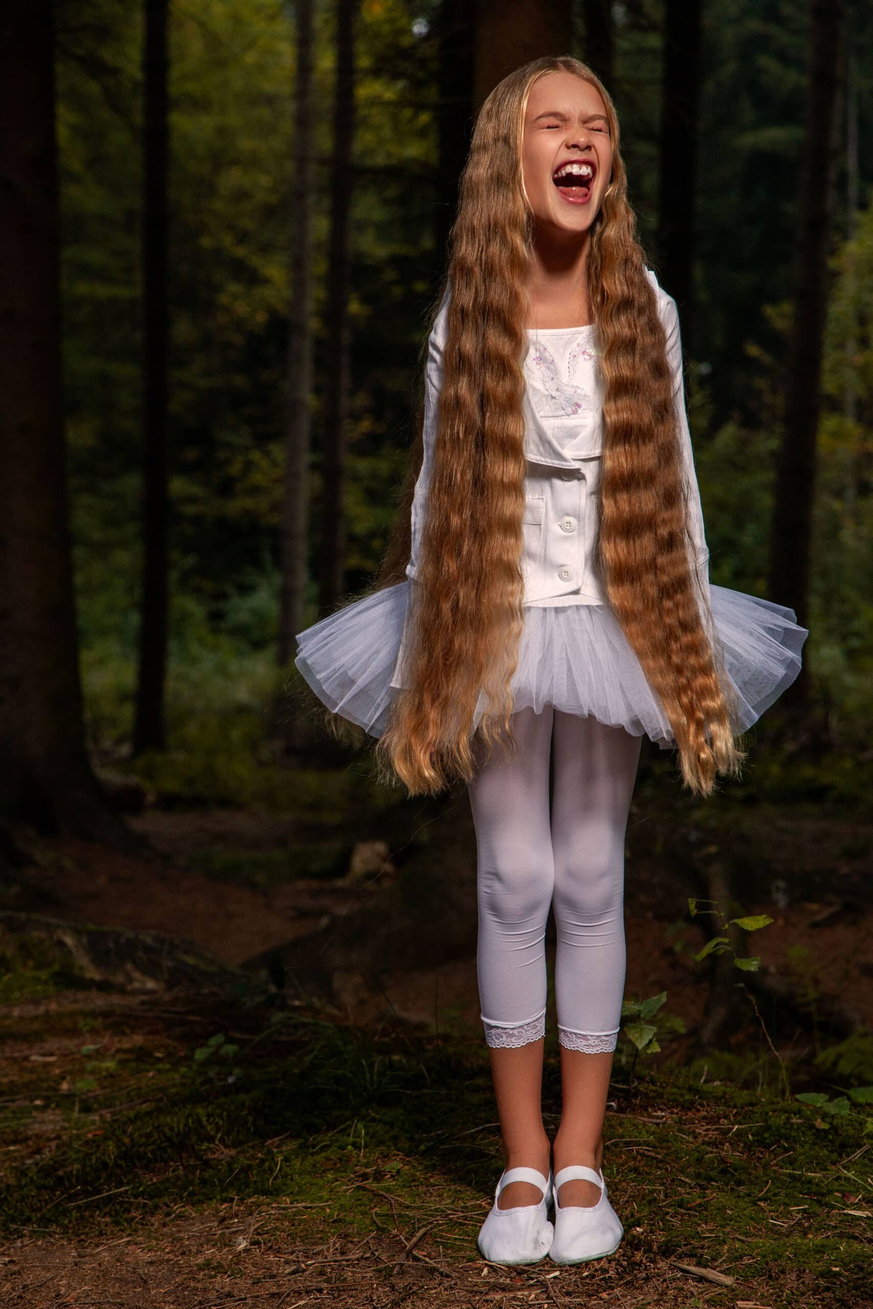 Ein junges Mädchen mit langen blonden Haaren lacht fröhlich in einem Wald. Es trägt eine weiße Bluse, ein Tutu und Leggings und wird von sanftem Licht angestrahlt. © Fotografie Tomas Rodriguez