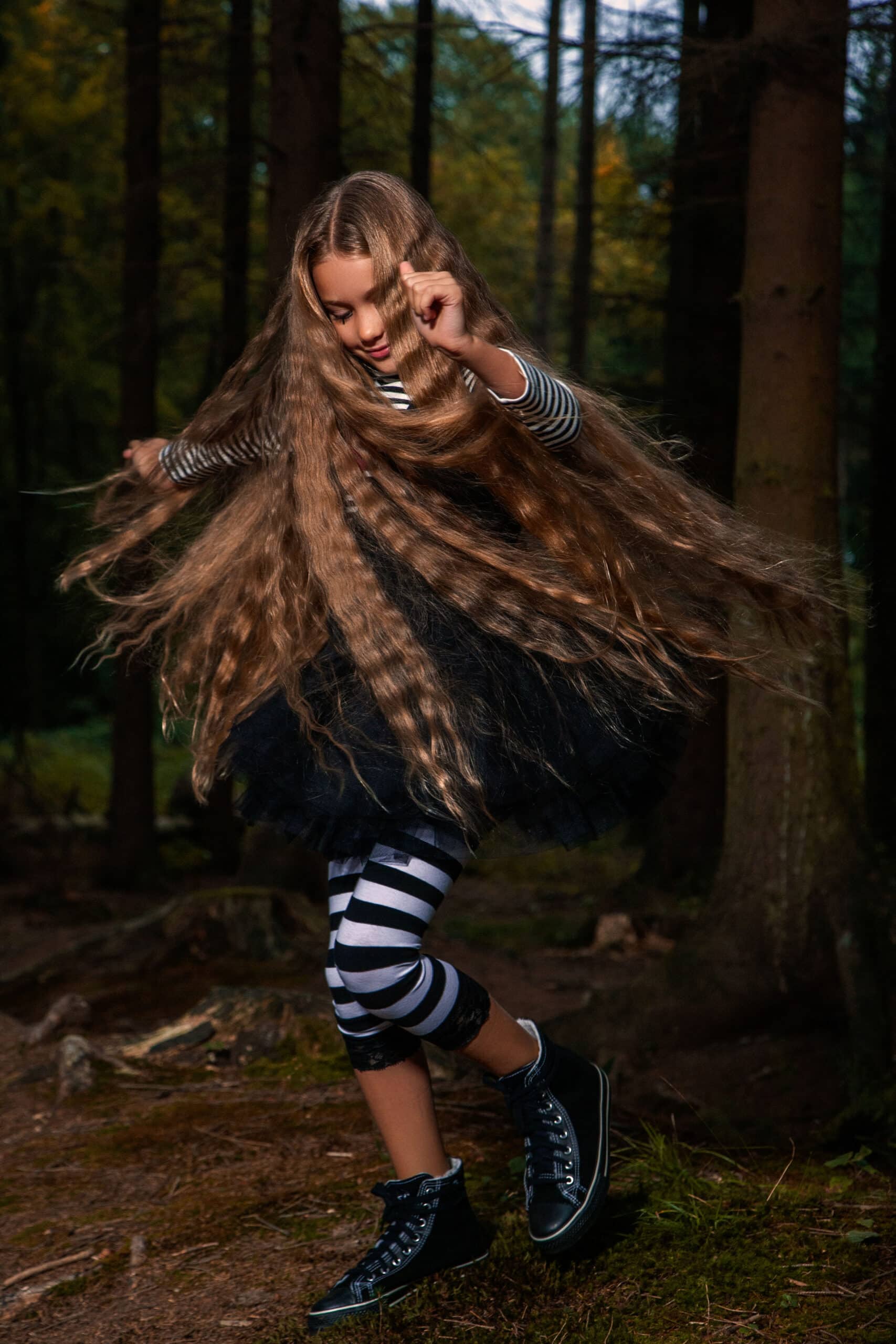 Ein junges Mädchen mit langem, wallendem Haar wirbelt fröhlich in einer Waldkulisse herum. Es trägt ein schwarzes Outfit und gestreifte Leggings. Ihre dynamische Bewegung betont ihr fächerartig ausgebreitetes Haar. © Fotografie Tomas Rodriguez