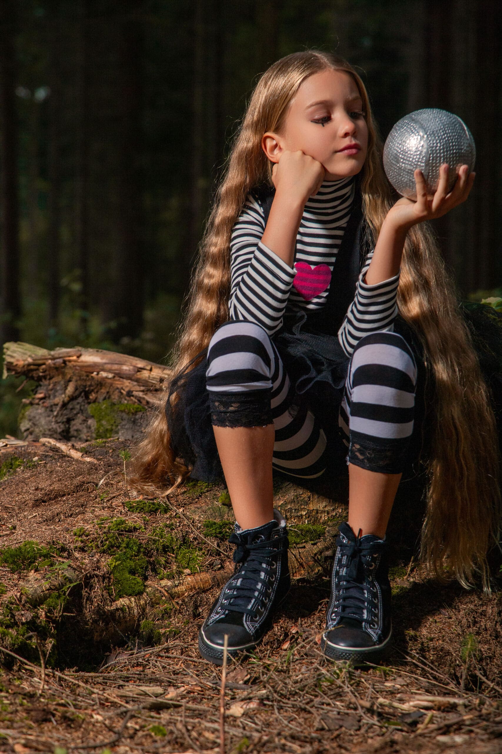 Ein junges Mädchen mit langen Haaren sitzt auf einem Baumstamm in einem Wald und blickt nachdenklich auf eine glänzende Discokugel, die sie in den Händen hält. Sie trägt ein gestreiftes Outfit mit passenden Knieschützern und High-Top-Sneakers. © Fotografie Tomas Rodriguez