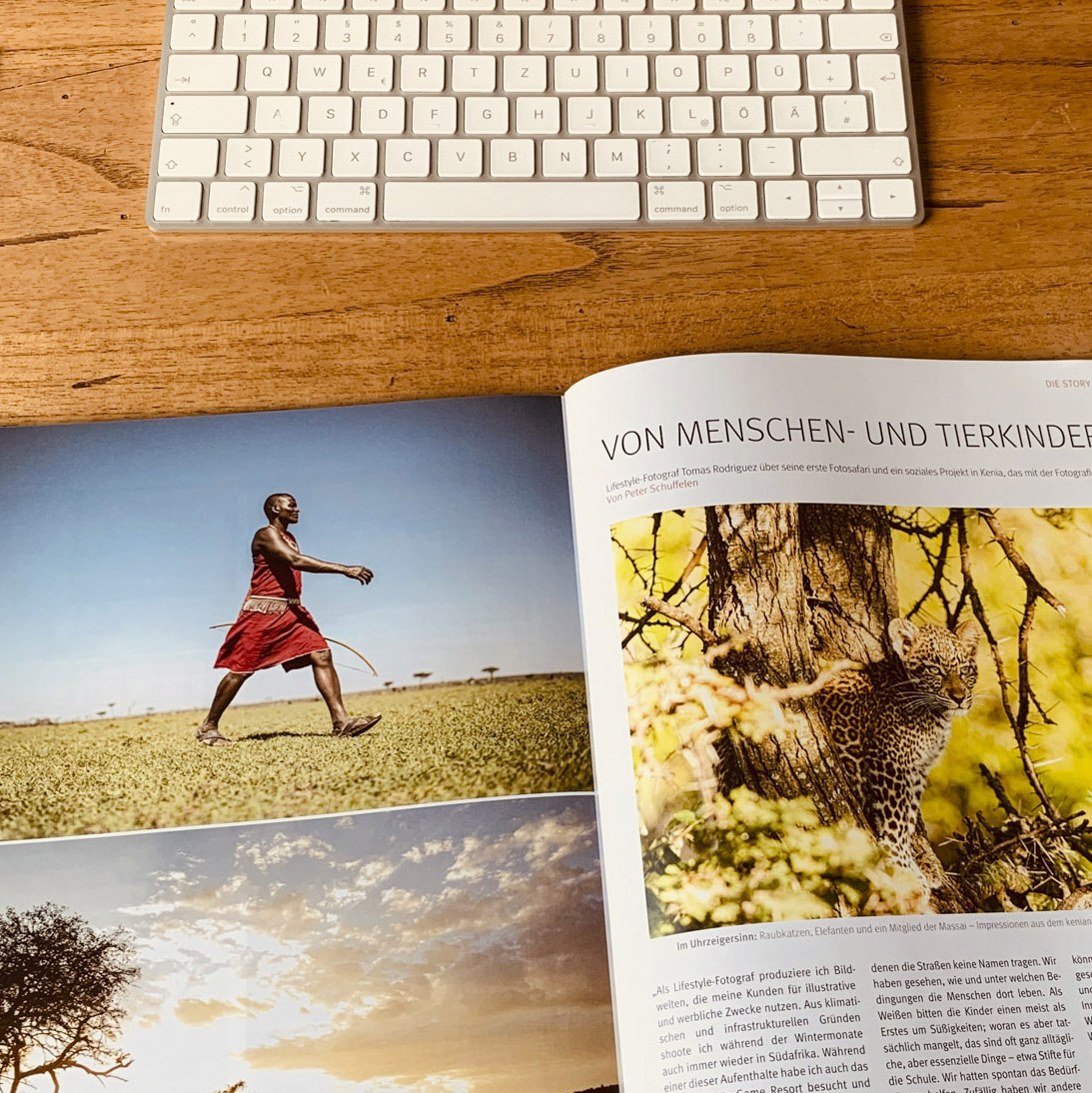 Eine aufgeschlagene Zeitschrift auf einem Holztisch zeigt das ausgedruckte Foto eines Mannes in traditioneller Kleidung, der durch eine Savanne läuft, neben der Tastatur eines Computers, was eine Arbeits- oder Lernumgebung suggeriert. © Fotografie Tomas Rodriguez