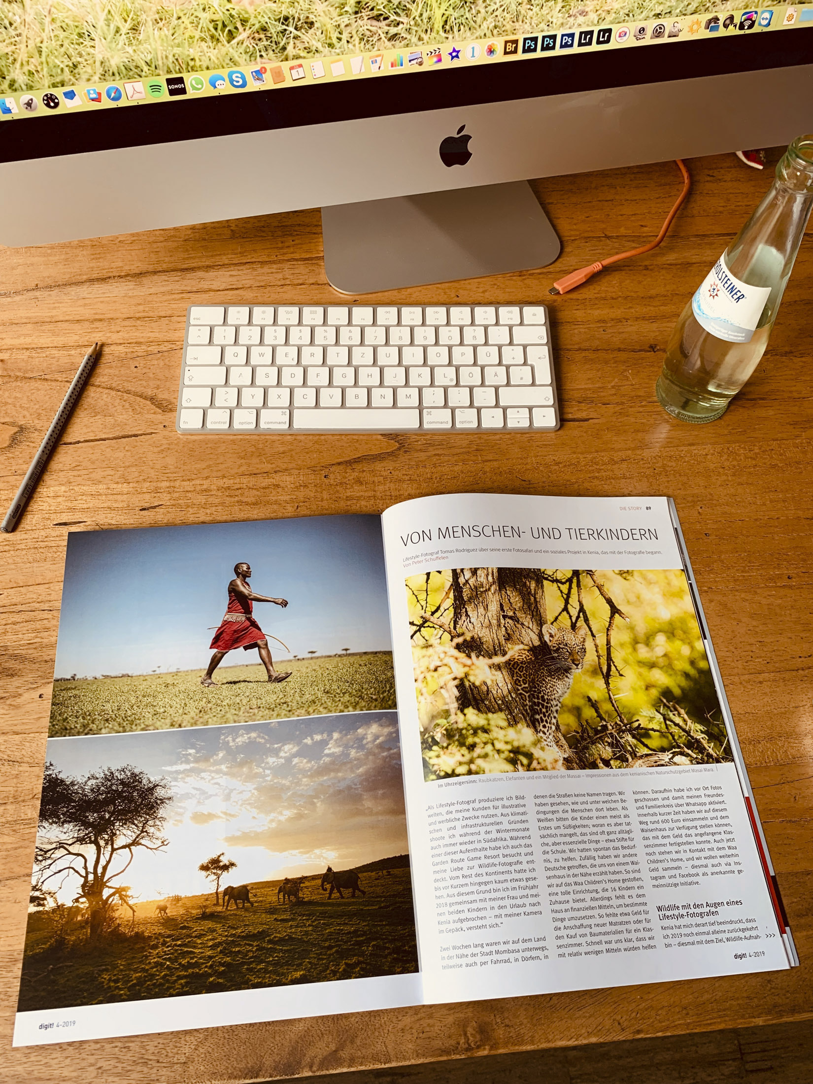 Ein gut organisierter Schreibtisch mit einem iMac, einer Tastatur und einem Magazin mit Naturfotografie, das auf einer Seite über Menschen und Tiere aufgeschlagen ist. Ein Bleistift und eine Flasche liegen ebenfalls auf dem Schreibtisch. © Fotografie Tomas Rodriguez