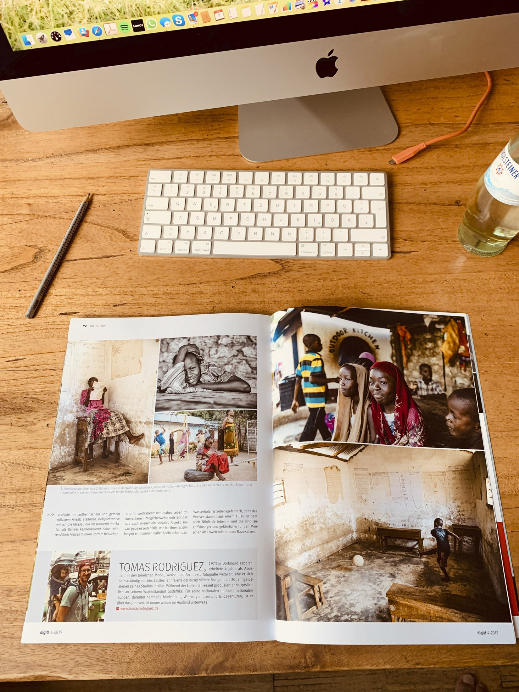 Ein Schreibtischarbeitsplatz mit einer aufgeschlagenen Zeitschrift mit fotojournalistischen Bildern auf einem Holztisch und einem Apple-Computer im Hintergrund. © Fotografie Tomas Rodriguez