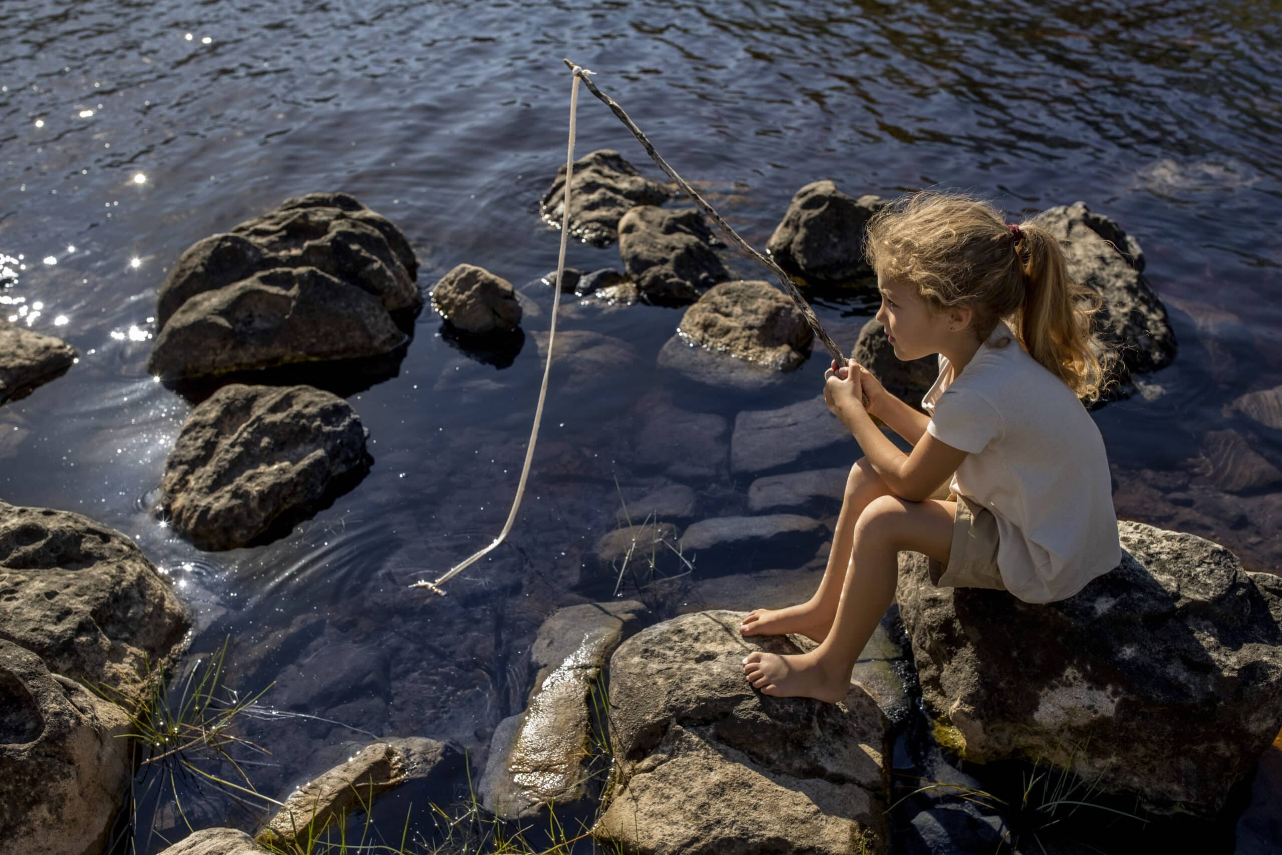 Ein junges Mädchen mit hellbraunem Haar und einem Pferdeschwanz sitzt barfuß auf einem Felsen am Ufer eines Flusses und hält eine Angelrute aus einem Stock in der Hand. Die Sonne spiegelt sich im Wasser und große Felsen sind im und um den Fluss verstreut. © Fotografie Tomas Rodriguez