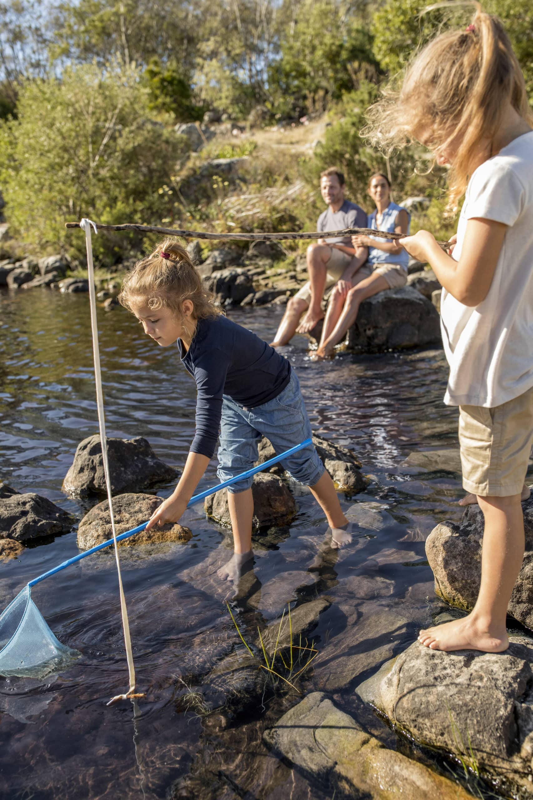 Zwei junge Mädchen spielen in einem flachen, felsigen Bereich eines Flusses. Ein Mädchen fängt mit einem Netz etwas aus dem Wasser, während das andere zusieht. Im Hintergrund sitzen ein Mann und eine Frau auf den Felsen und beobachten die Kinder. Bäume und Grünflächen sind zu sehen. © Fotografie Tomas Rodriguez