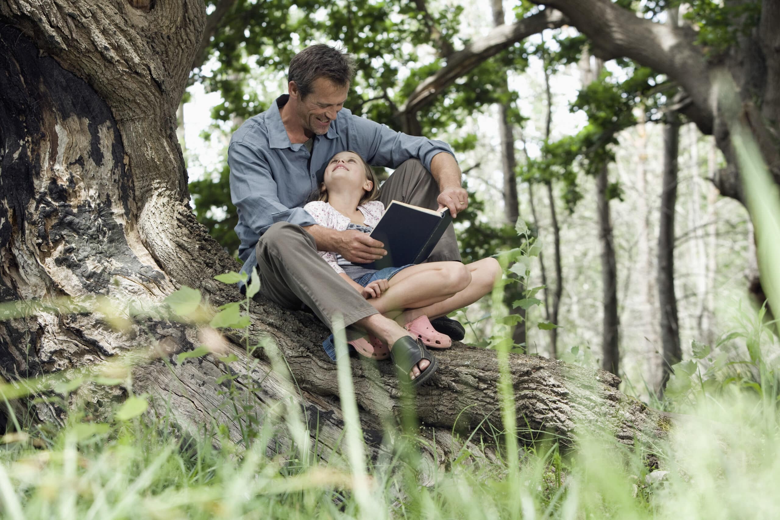 Ein Mann sitzt auf einem Ast und liest ein Buch. Ein junges Mädchen sitzt auf seinem Schoß. Sie sind draußen, umgeben von einem Wald mit grünem Laub. Der Mann sieht das Mädchen an, das ihn anlächelt. © Fotografie Tomas Rodriguez