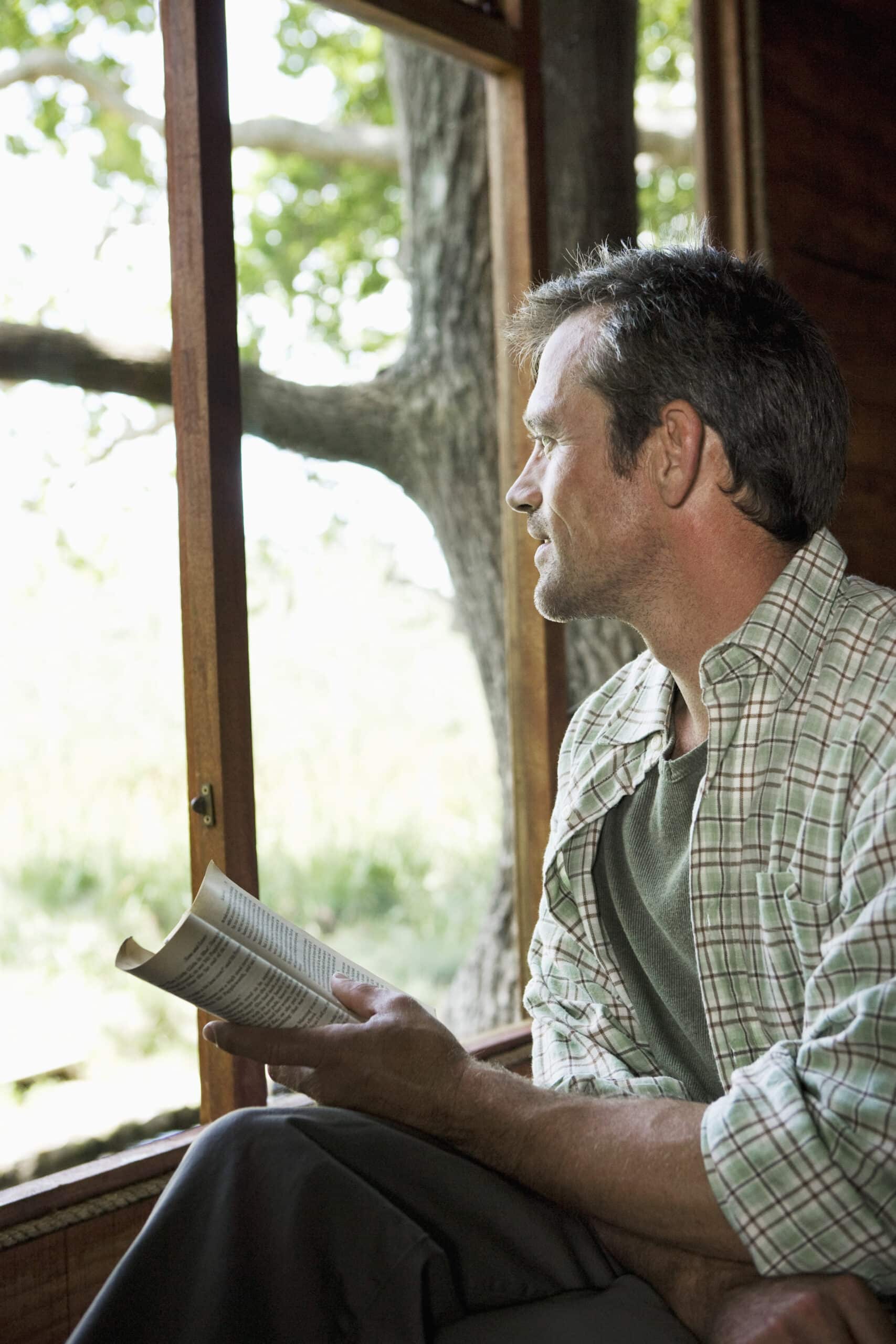 Ein Mann mit kurzen dunklen Haaren, einem grünen Hemd und einer karierten Jacke, hält ein offenes Buch in der Hand und blickt nachdenklich aus einem Holzfenster. Durch das Fenster sind ein großer Baum und viel Grün zu sehen, was eine friedliche, natürliche Umgebung suggeriert. © Fotografie Tomas Rodriguez