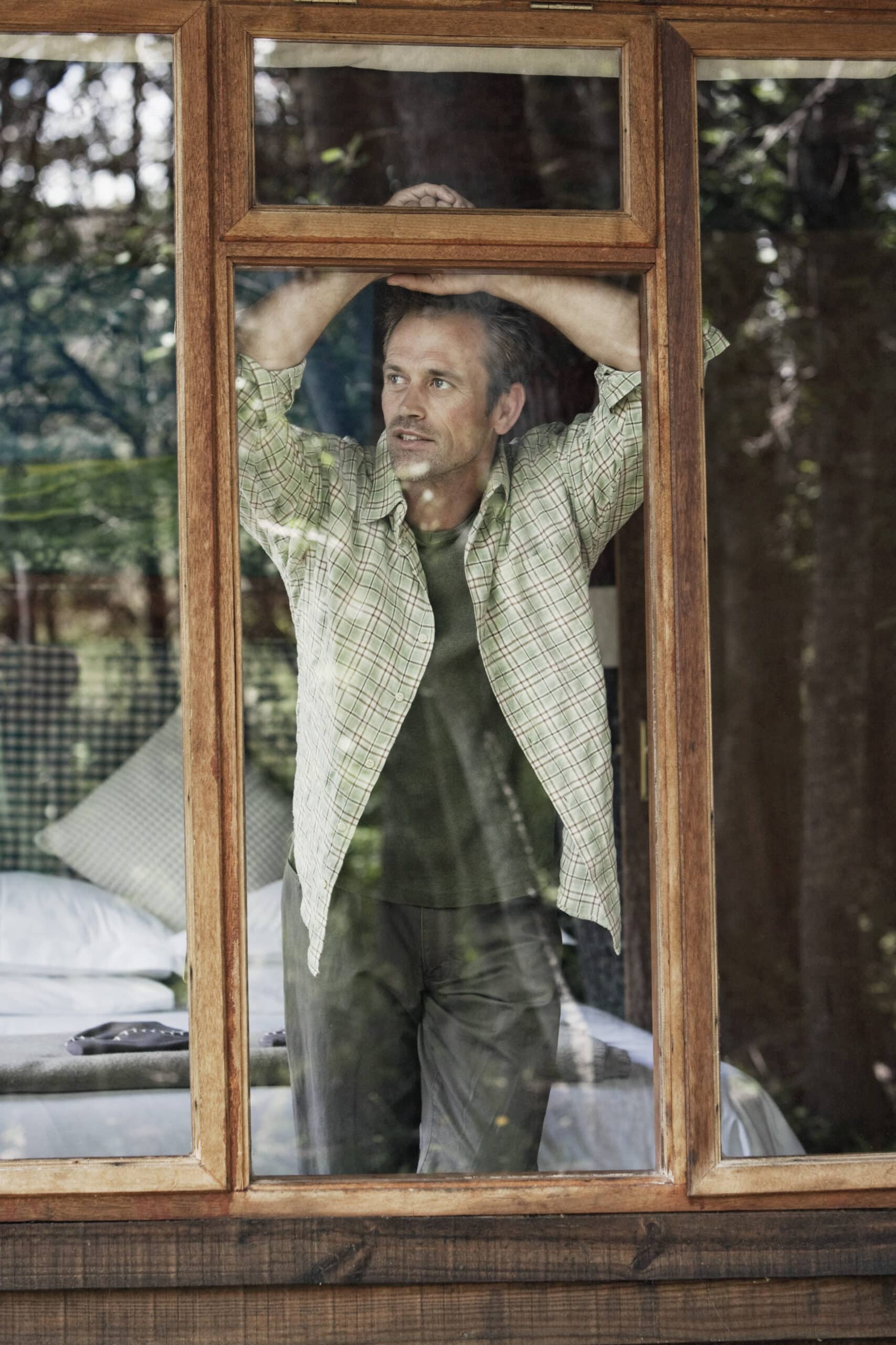Ein Mann in einem grün karierten Hemd und dunkler Hose steht drinnen und schaut aus einem Fenster mit Holzrahmen. Er hat seine Arme erhoben und berührt die Oberkante des Fensterrahmens. Im Hintergrund ist ein Bett mit Kissen zu sehen, und die Szene draußen zeigt Bäume und Grün. © Fotografie Tomas Rodriguez