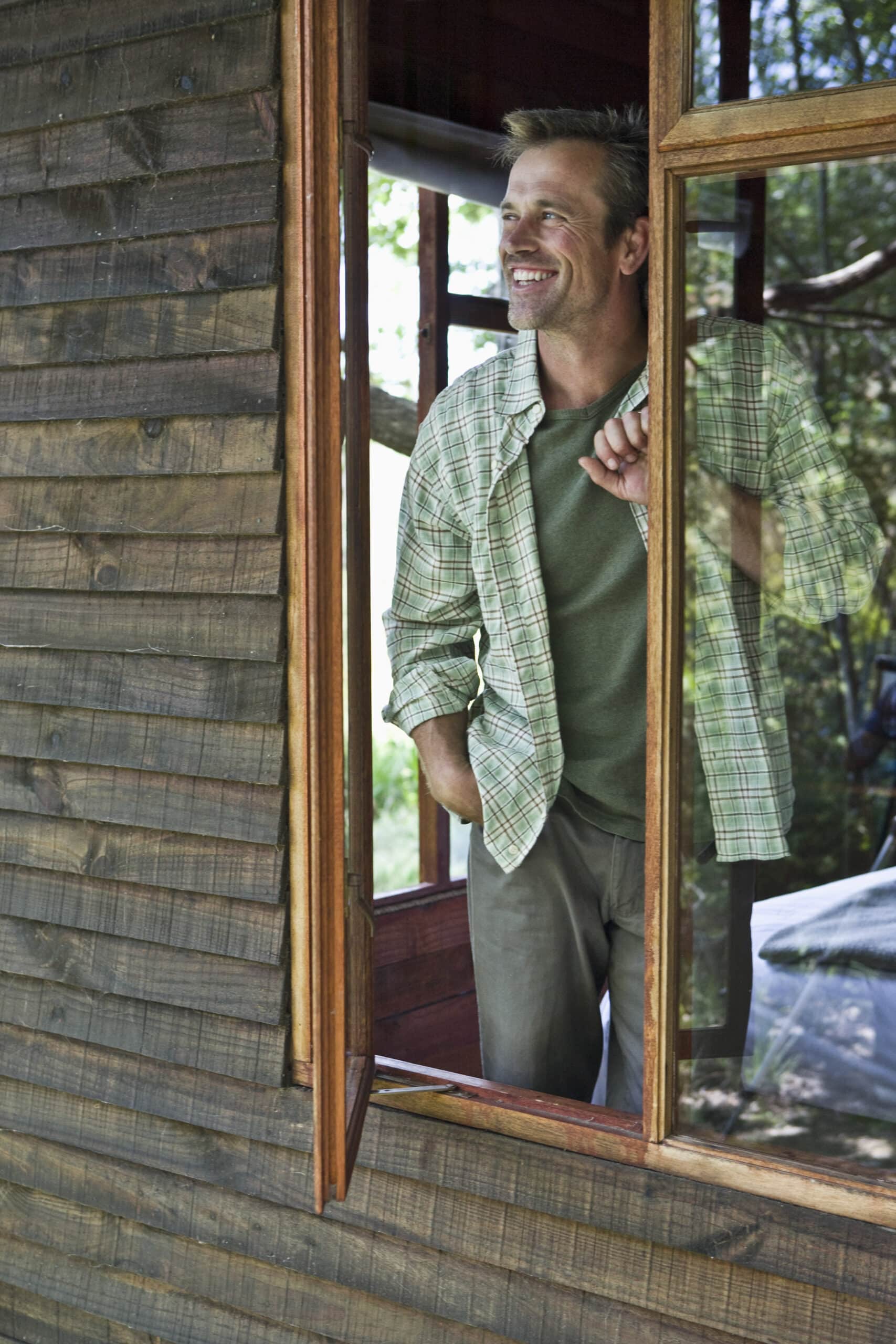 Ein Mann mit kurzen Haaren und Bart, der ein grün kariertes Hemd und Khakihosen trägt, steht am offenen Fenster einer Holzhütte. Er lächelt und schaut nach draußen, eine Hand ruht auf dem Fensterrahmen. Im Hintergrund sind Bäume zu sehen. © Fotografie Tomas Rodriguez