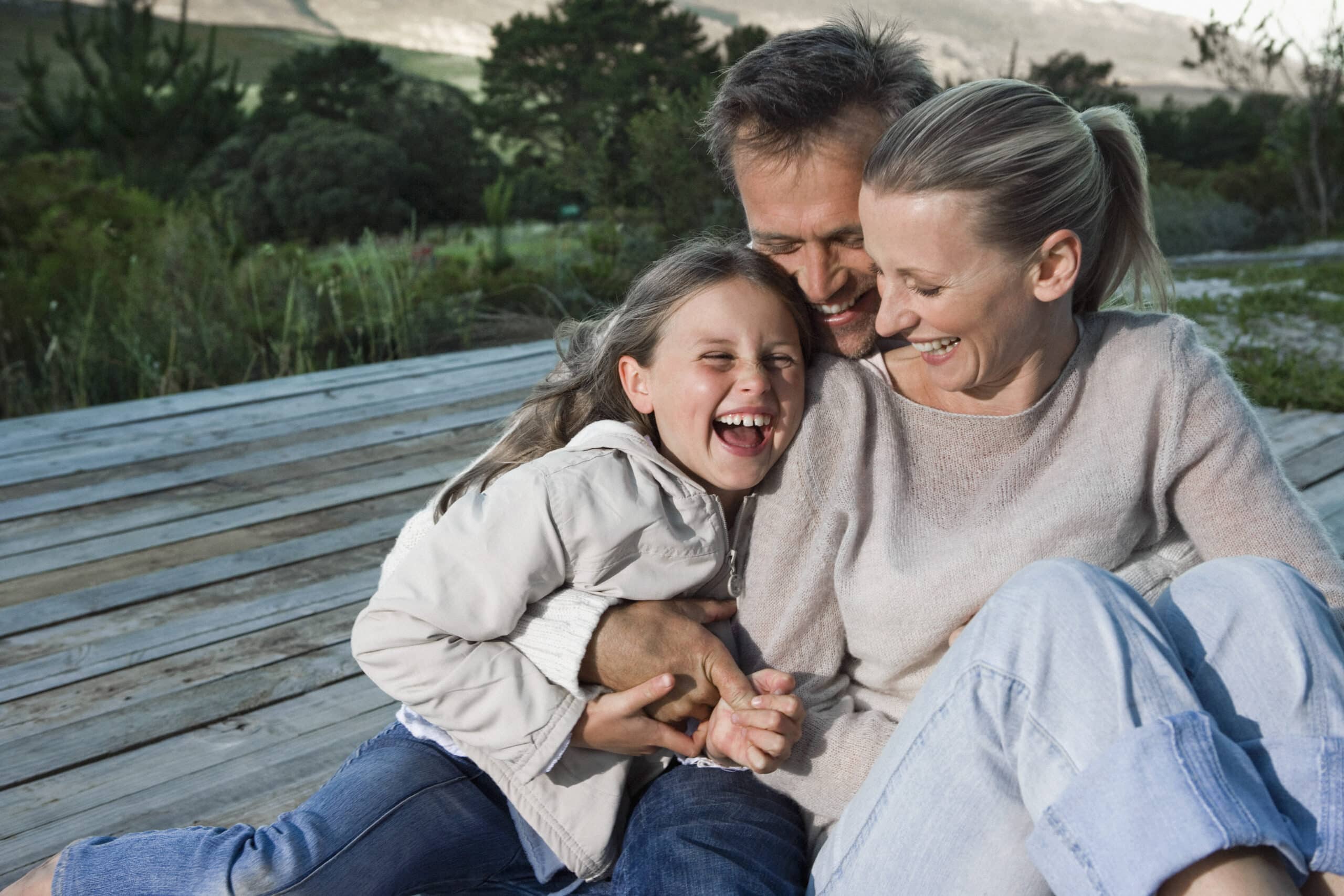 Eine dreiköpfige Familie, bestehend aus einem Mann, einer Frau und einem jungen Mädchen, sitzt auf einem Holzdeck. Sie drängen sich eng aneinander, lachen und lächeln vor einer malerischen Naturkulisse aus Bäumen und Hügeln. Das Mädchen in einer weißen Jacke sitzt auf dem Schoß des Mannes. © Fotografie Tomas Rodriguez