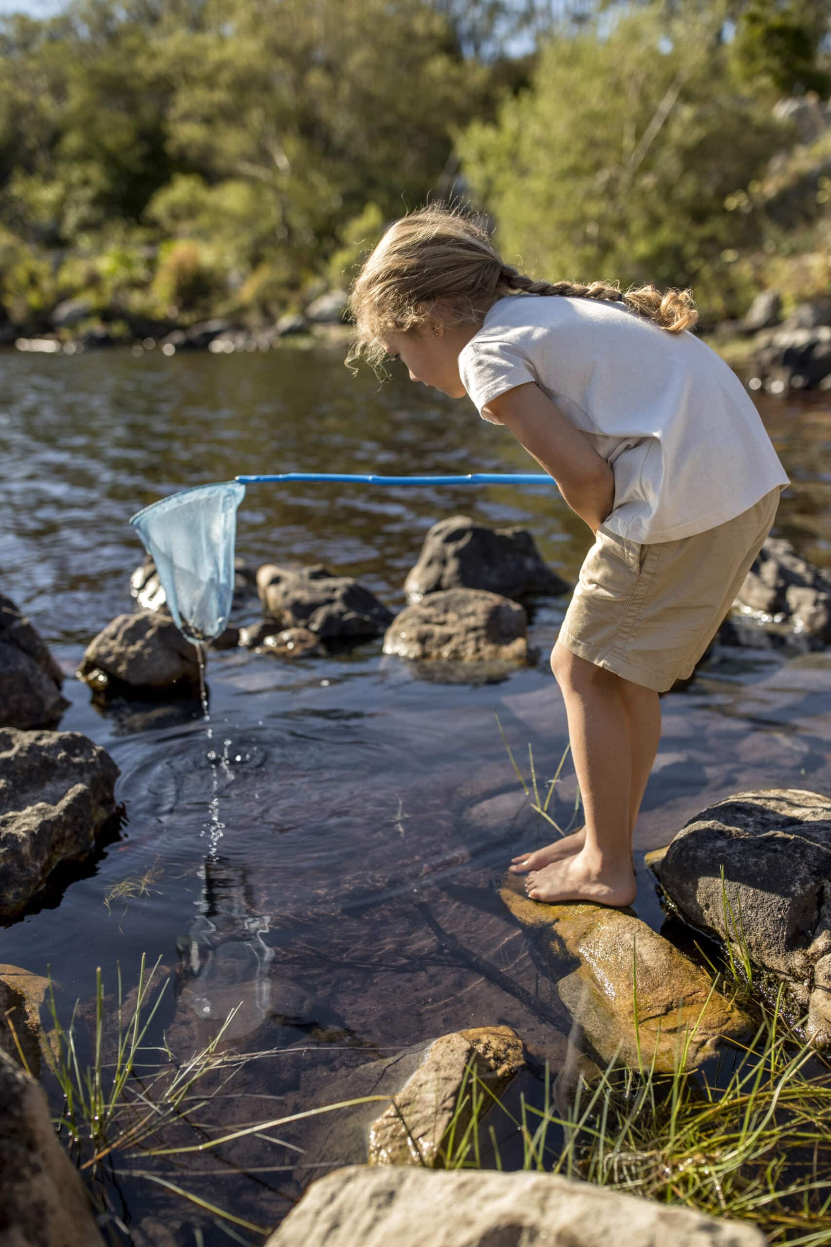 Ein kleines Kind mit geflochtenen Haaren steht barfuß auf Felsen am Rande eines Baches und hält ein blaues Netz ins Wasser. Es trägt ein weißes Hemd und beige Shorts. Im Hintergrund sind Grünpflanzen und Bäume zu sehen. Die Szene ist hell und sonnig. © Fotografie Tomas Rodriguez