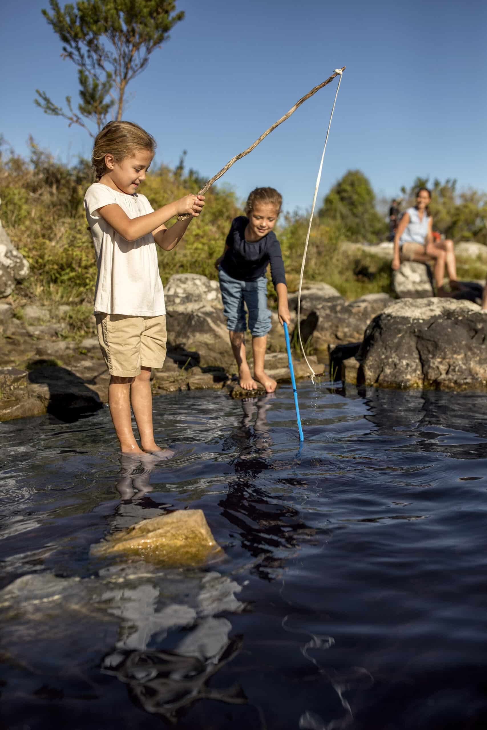 Zwei Kinder angeln mit improvisierten Angelruten an einem Teich. Ein Kind überprüft die Angelschnur, während das andere seine Angelrute ins Wasser wirft. Ein Erwachsener sitzt im Hintergrund auf einem Felsen und beobachtet die Szene. Das Wetter ist sonnig und der Himmel klar. © Fotografie Tomas Rodriguez