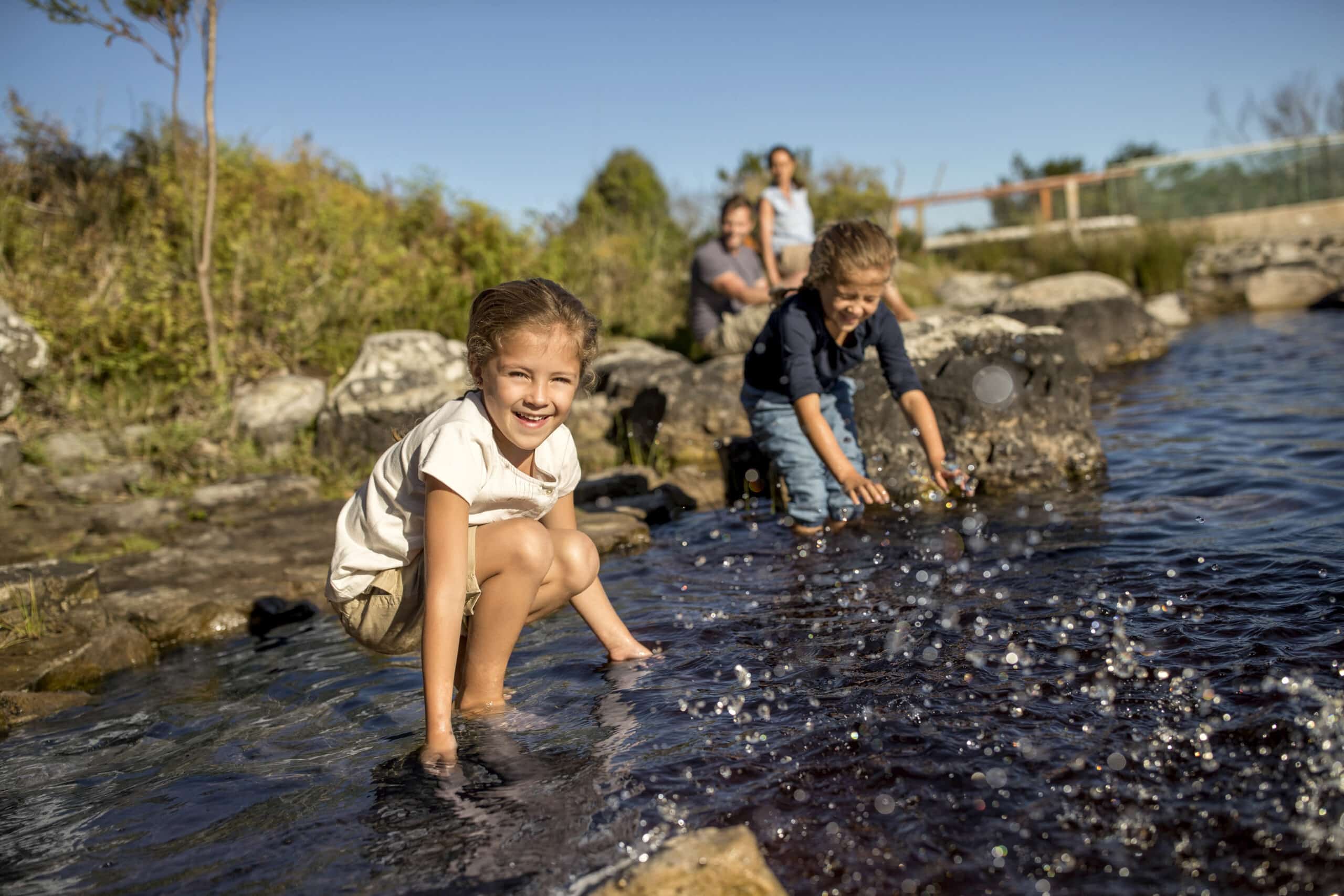 Zwei Kinder spielen in einem seichten Bach und spritzen mit ihren Händen Wasser. Ein kleines Mädchen in weißem Hemd und Shorts lächelt in die Kamera, während ein Junge in dunklem Hemd auf das Wasser blickt. Zwei Erwachsene stehen im Hintergrund bei Bäumen und Felsen und beobachten die Kinder. © Fotografie Tomas Rodriguez
