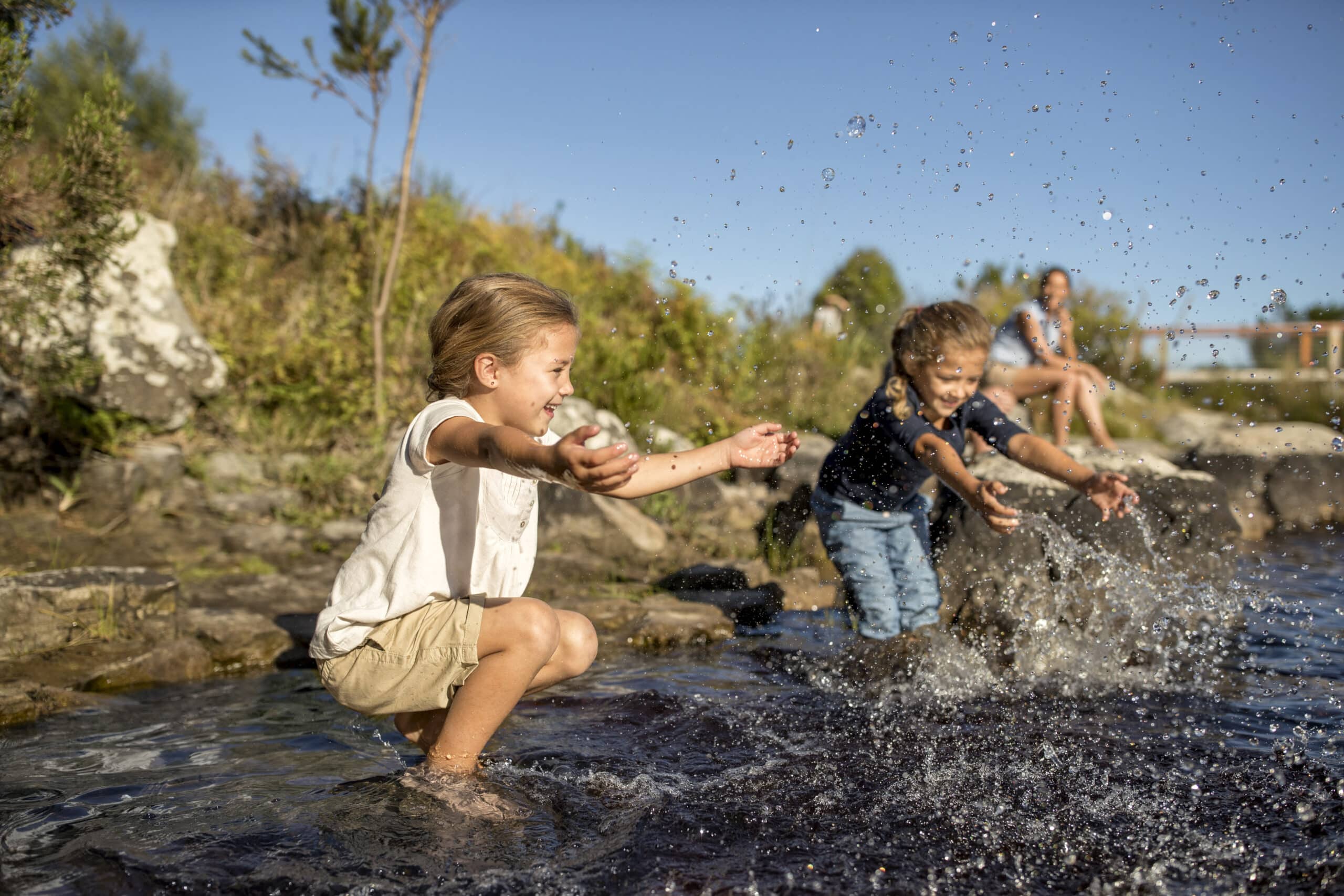 Zwei Kinder bespritzen sich gegenseitig spielerisch mit Wasser, während sie in einem seichten, steinigen Bach stehen. Ein Kind hockt mit ausgestreckten Armen, das andere steht ihnen gegenüber, beide lächeln. Ein Erwachsener sitzt auf einem Felsen im Hintergrund, beobachtet und lächelt. Es ist ein sonniger Tag. © Fotografie Tomas Rodriguez